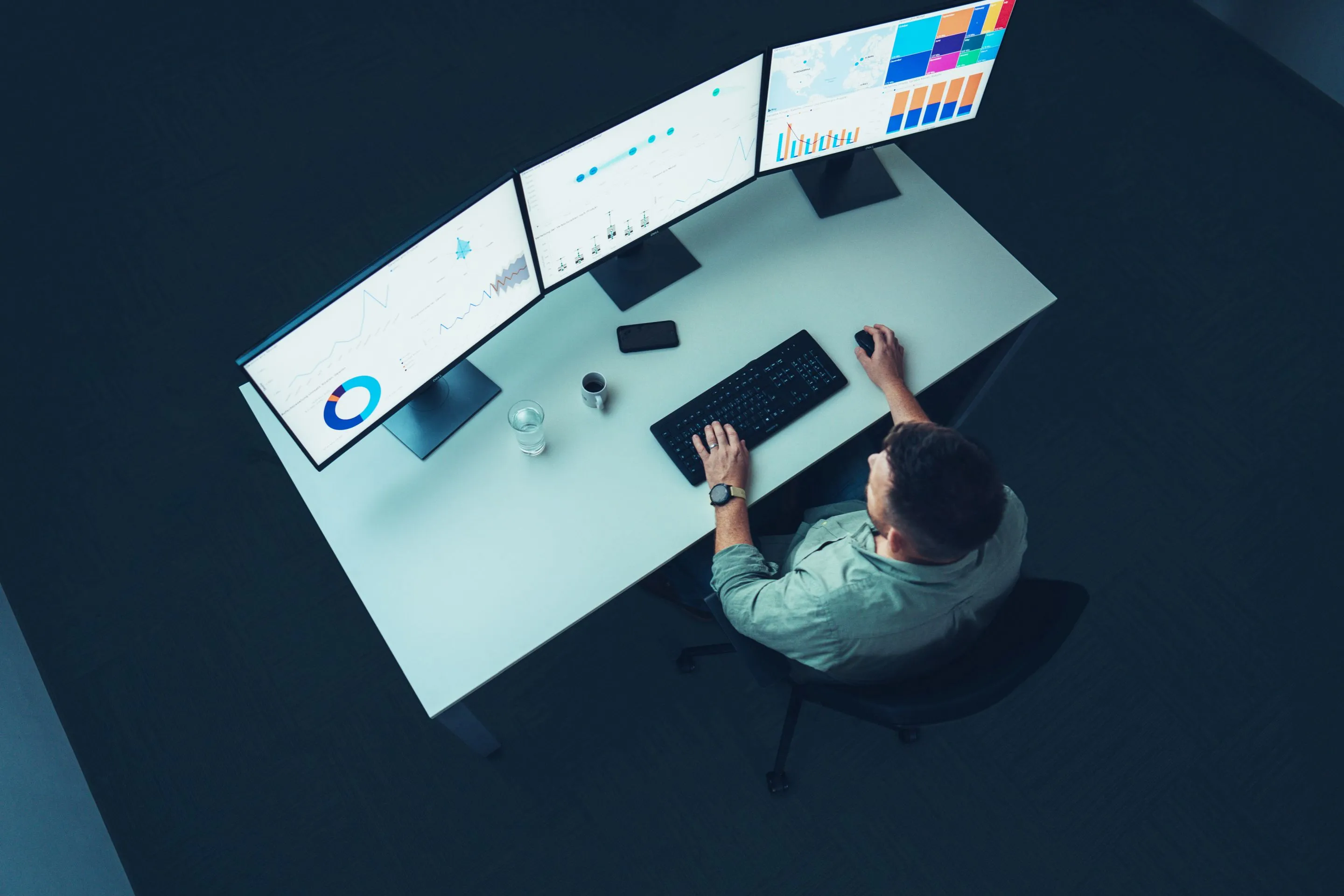 Person sitzt an einem Schreibtisch und arbeitet an einem Computer mit drei Monitoren, die verschiedene Diagramme und Datenvisualisierungen anzeigen.