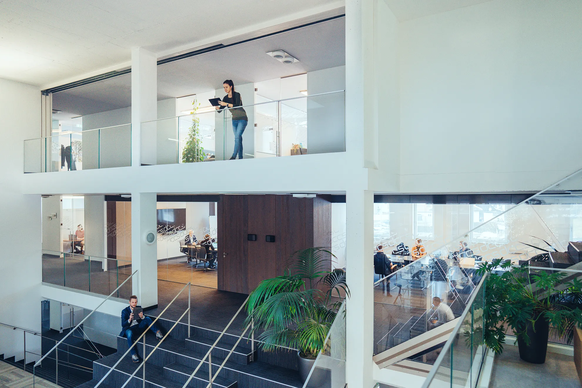 Modernes Bürogebäude mit Glaswänden, mehreren Personen, die an Tablets und Gesprächen beteiligt sind, und einer Treppe mit Pflanzen.