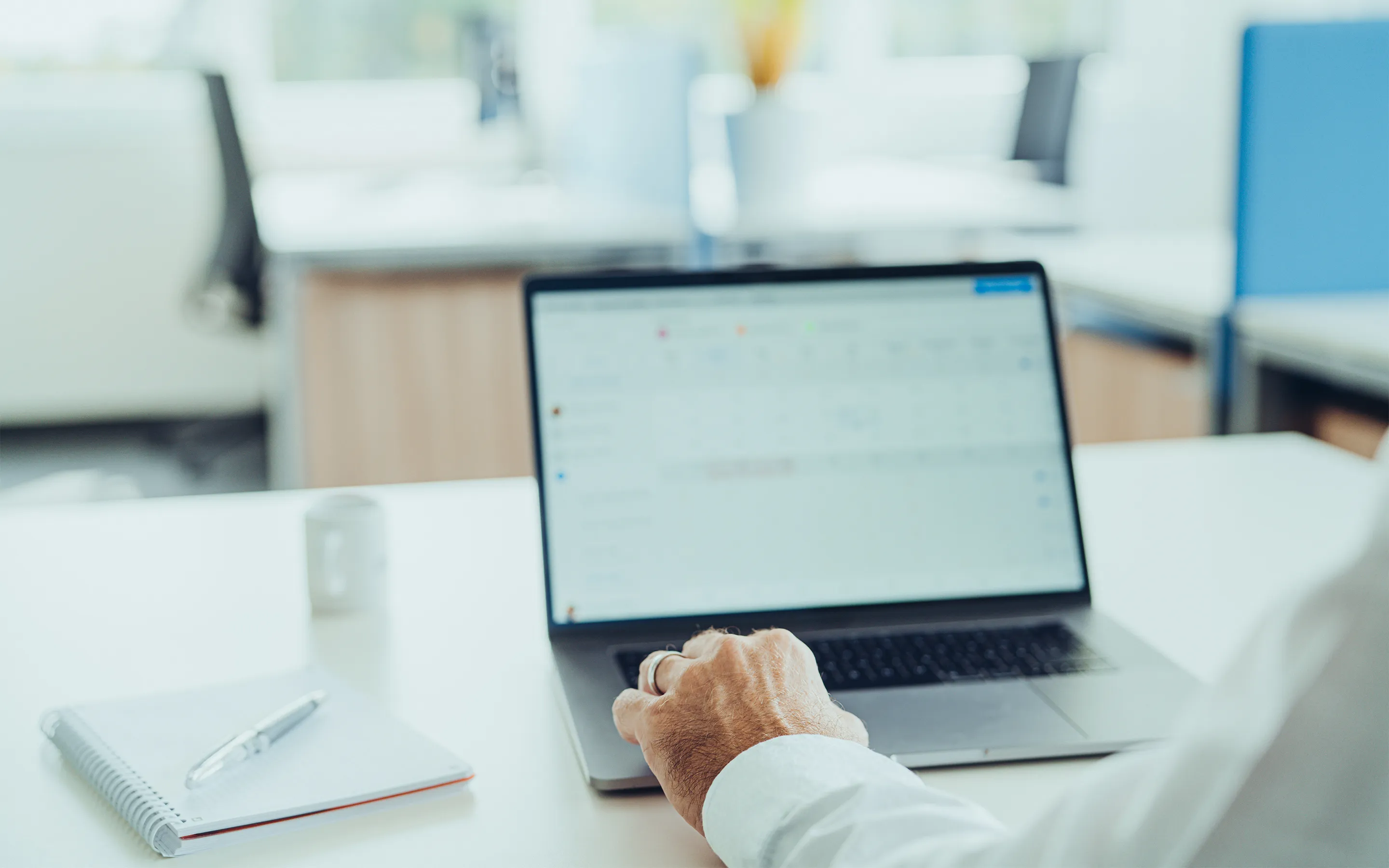 Hand eines Mannes in weißem Hemd, der auf einem Laptop mit Kalenderansicht in einem modernen Büro arbeitet.