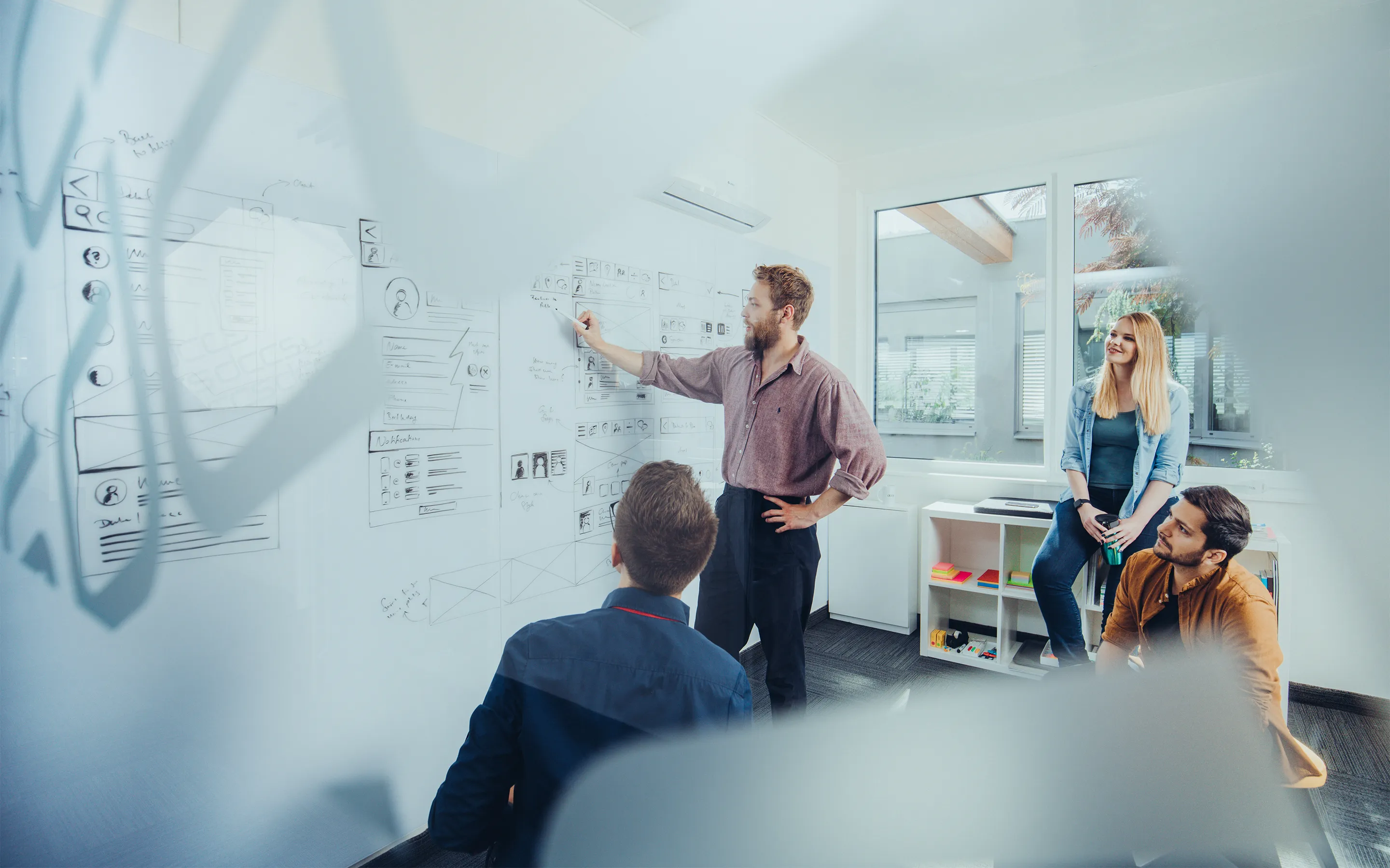 Vier Personen in einem hellen Büroraum diskutieren vor einer Glaswand mit UI/UX-Designskizzen.