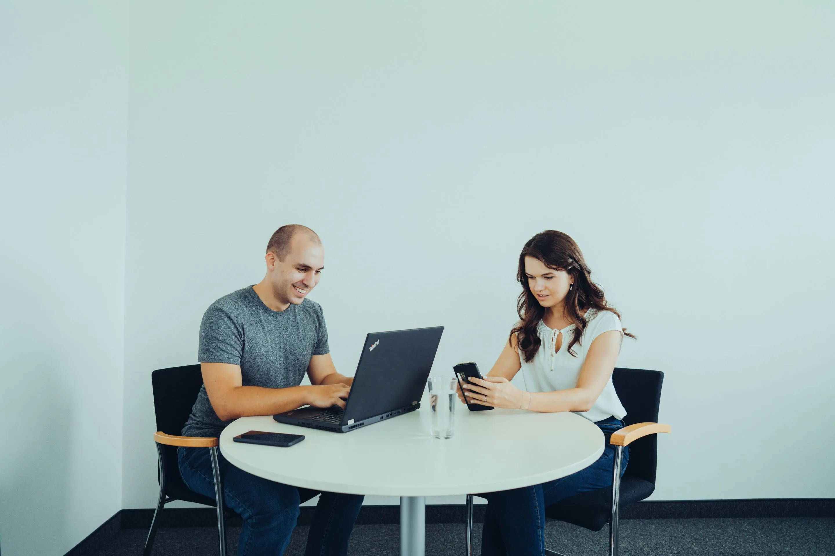 Ein Mann und eine Frau arbeiten an einem runden Tisch, der Mann nutzt einen Laptop und die Frau hält ein Smartphone.