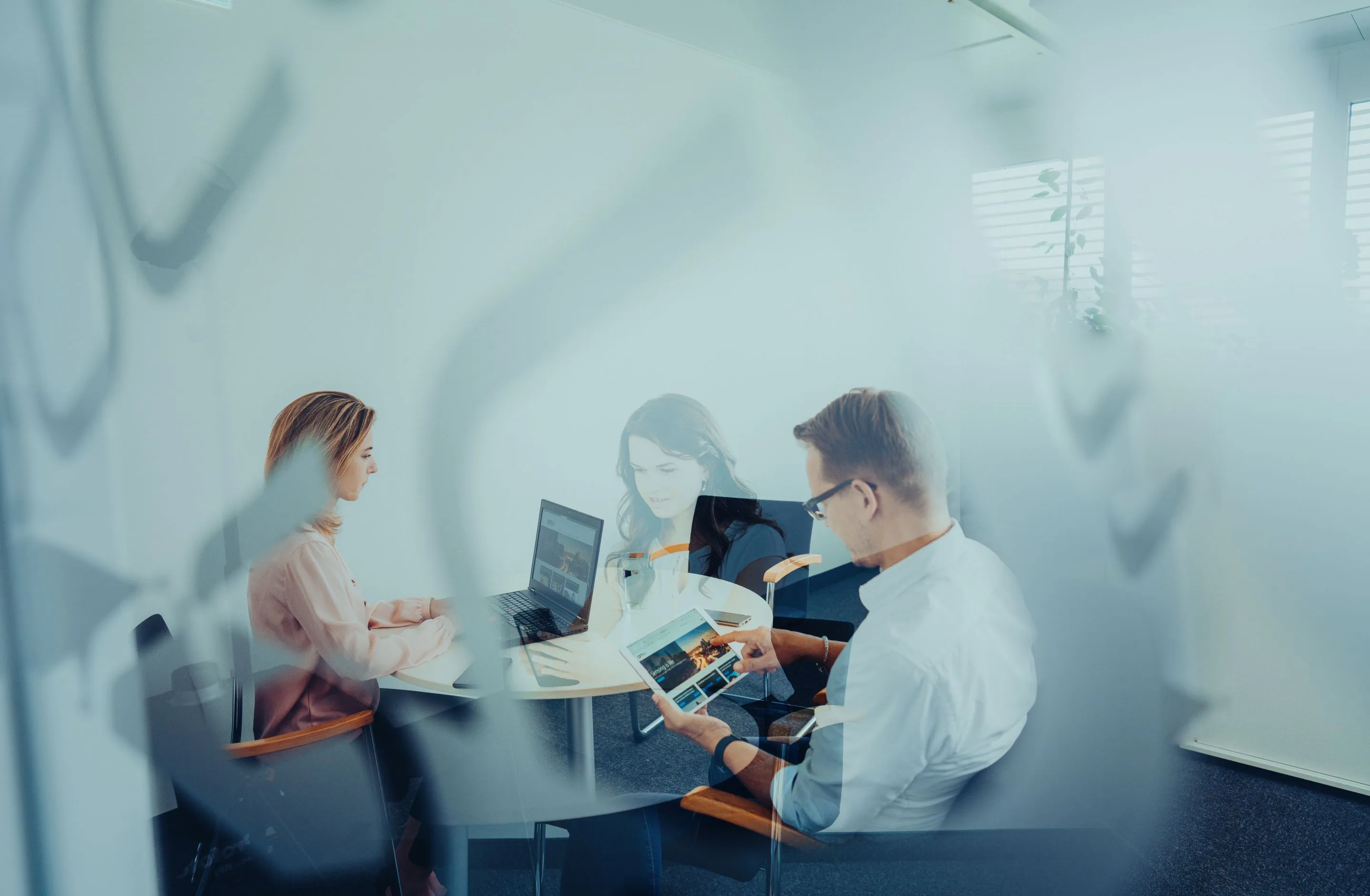 Drei Personen bei einer Besprechung in einem Büro, eine Frau arbeitet am Laptop, ein Mann nutzt ein Tablet.