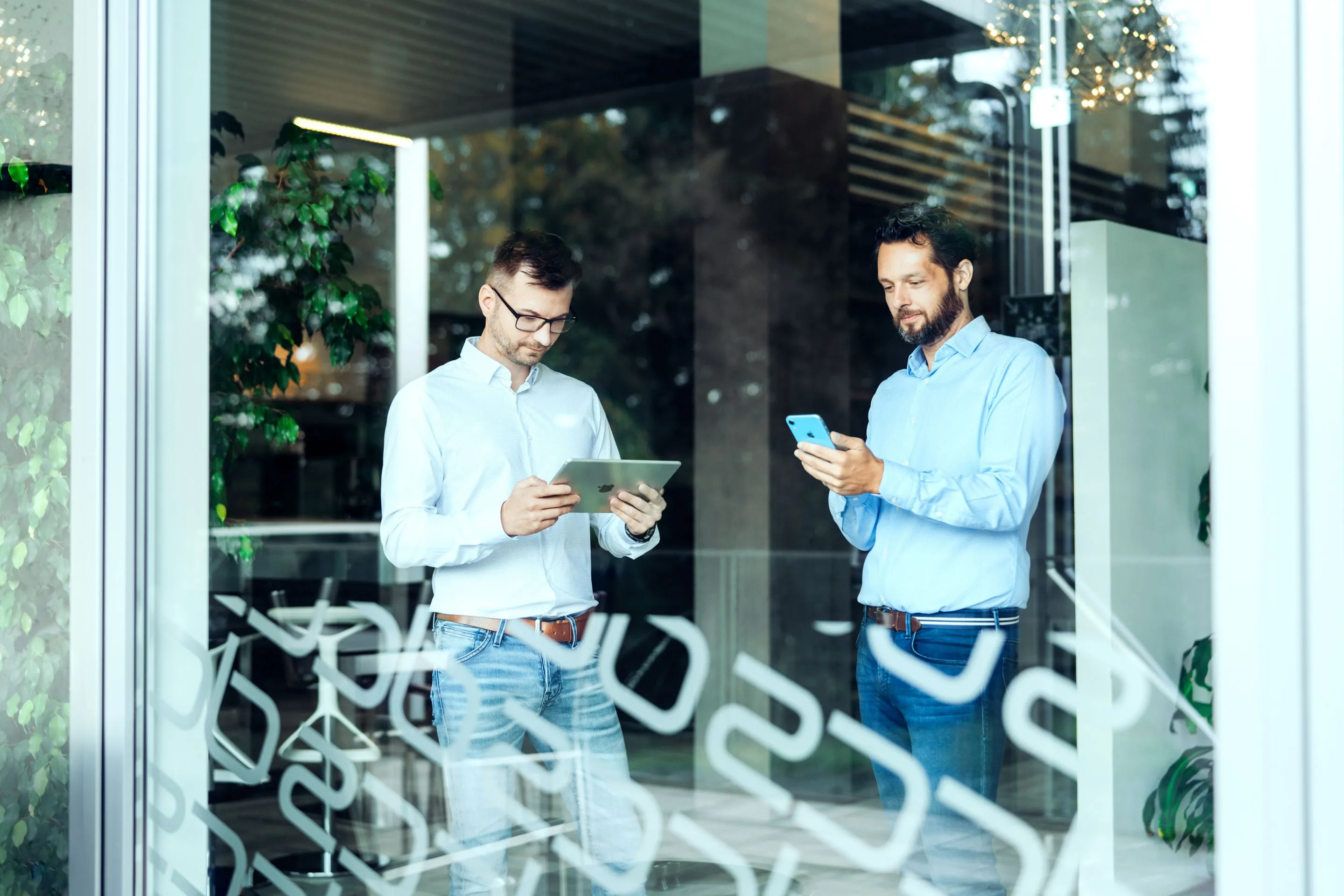 Zwei Männer in casual Businesskleidung nutzen ein Tablet und ein Smartphone in einem modernen Büro.
