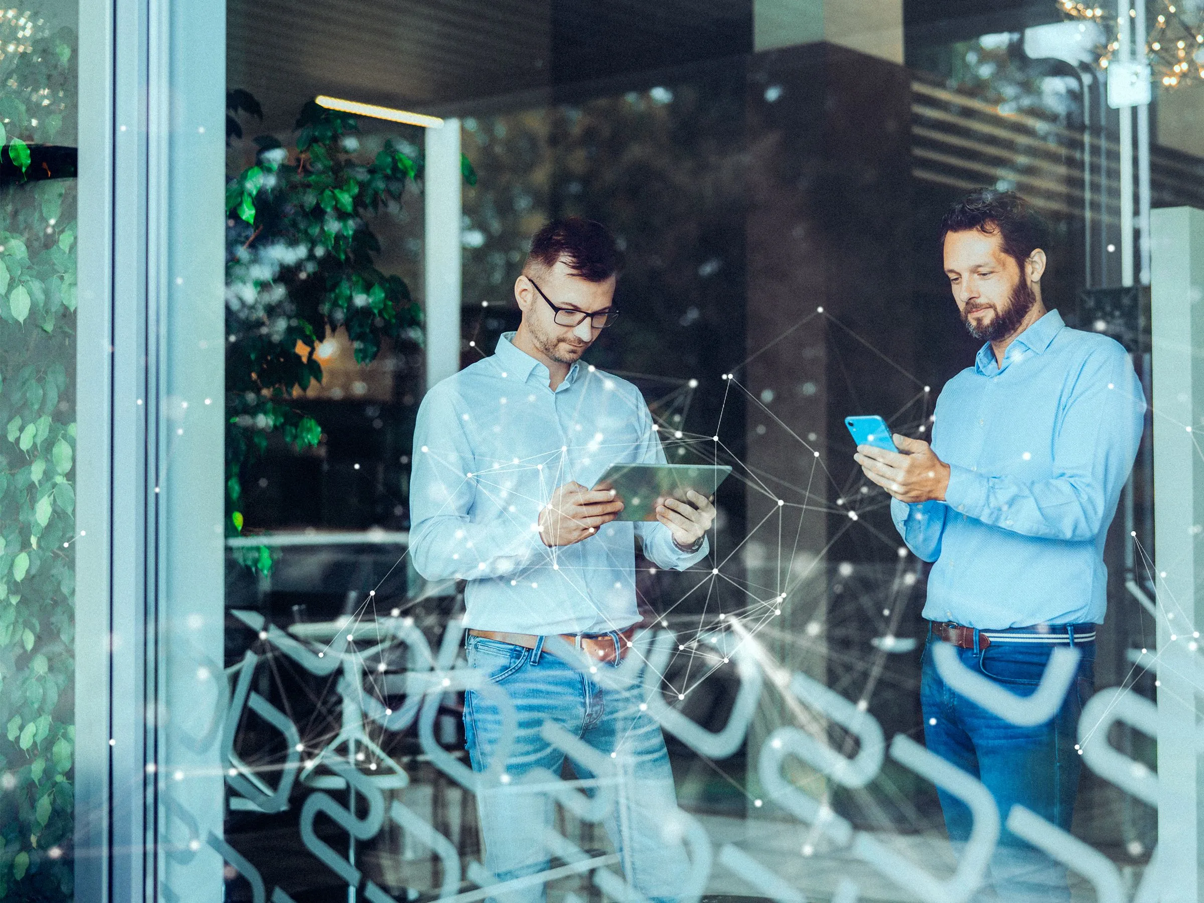 Zwei Männer in lässiger Geschäftskleidung verwenden ein Tablet und ein Smartphone in einem modernen Bürogebäude mit Glasfront.