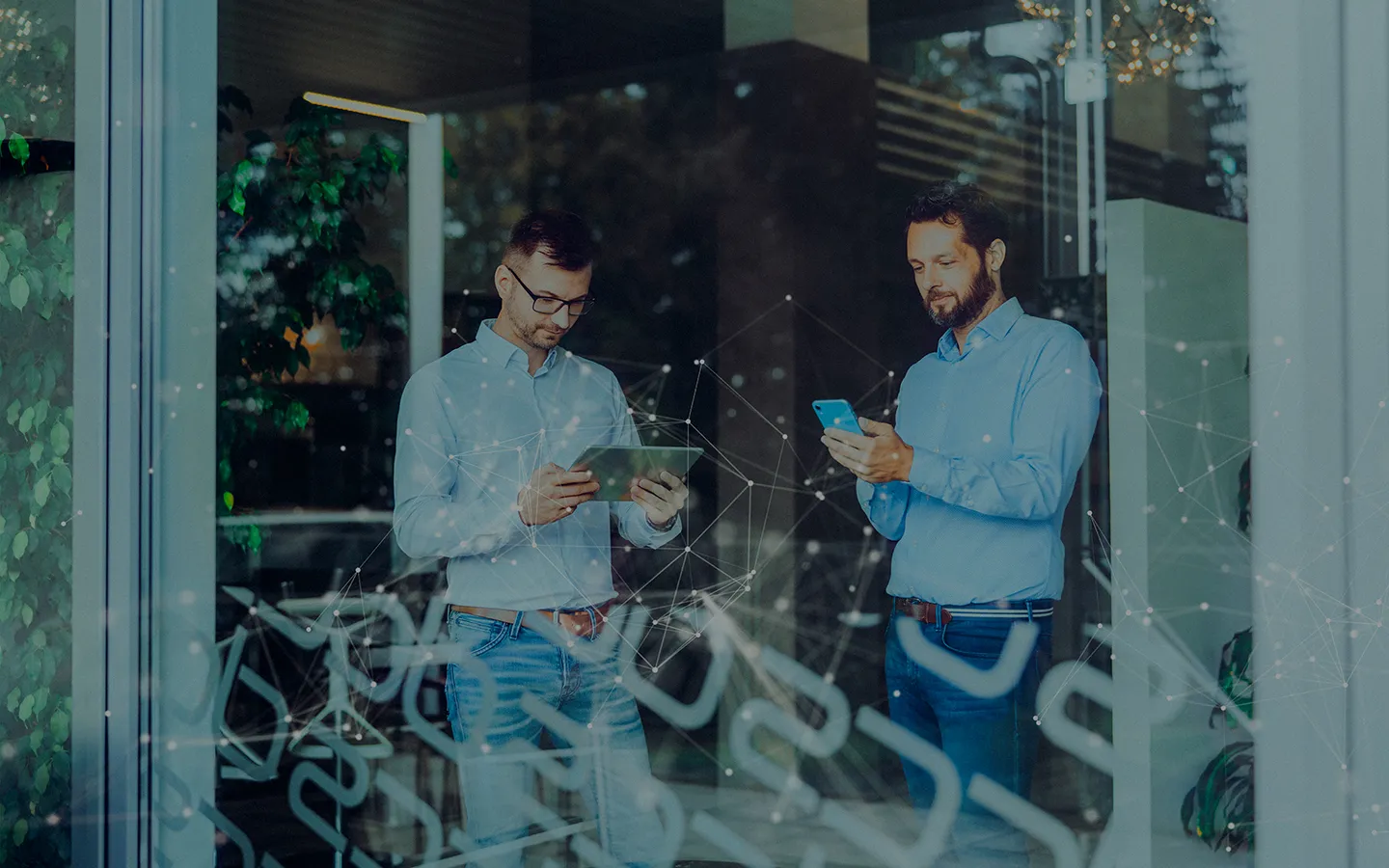 Zwei Männer in hellblauen Hemden stehen in einem modernen Büro und nutzen ein Tablet beziehungsweise ein Smartphone.