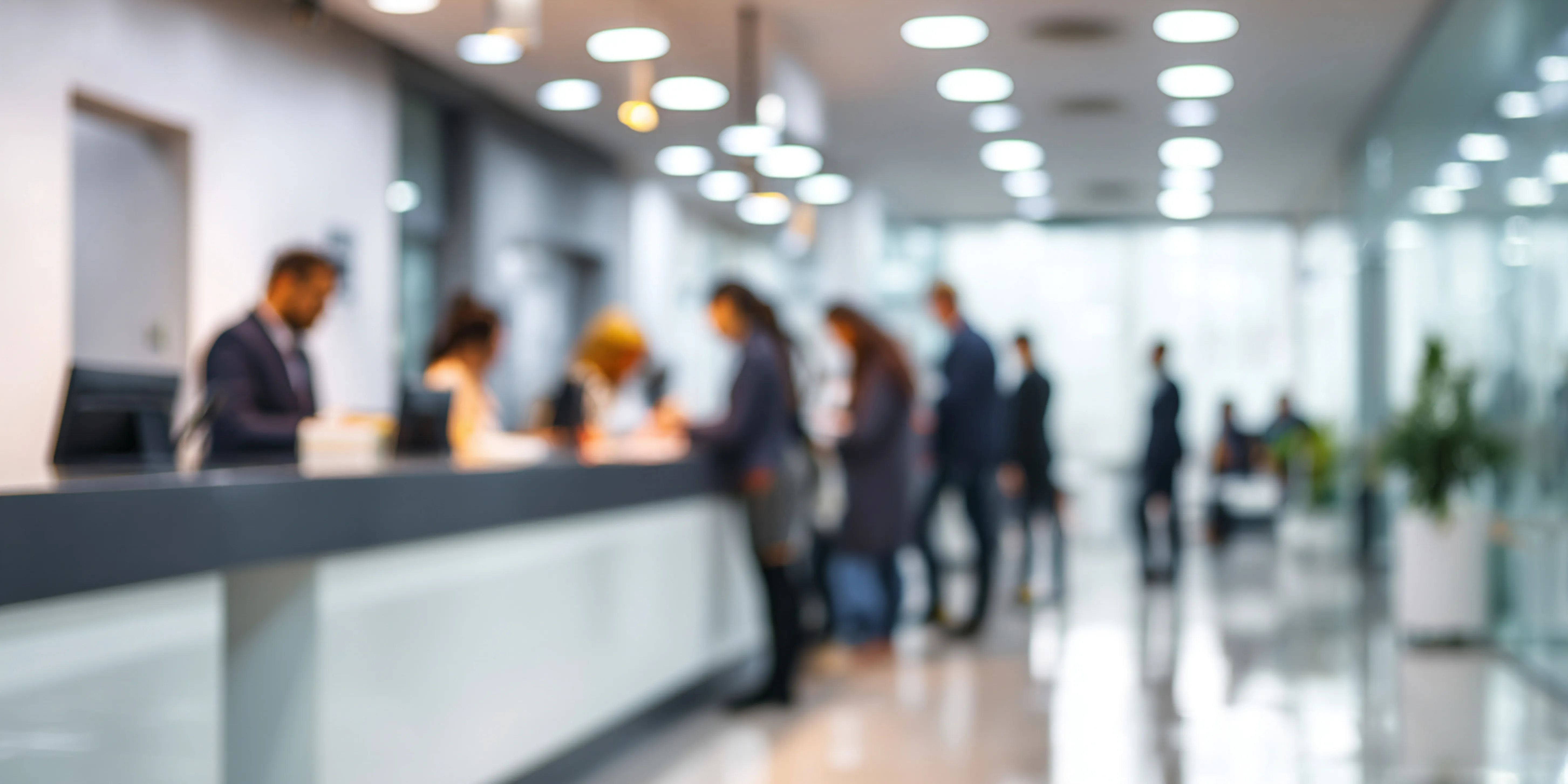 Verschwommener Blick auf Menschen in formeller Kleidung, die an einer Rezeption in einem modernen, hellen Bürogebäude stehen.