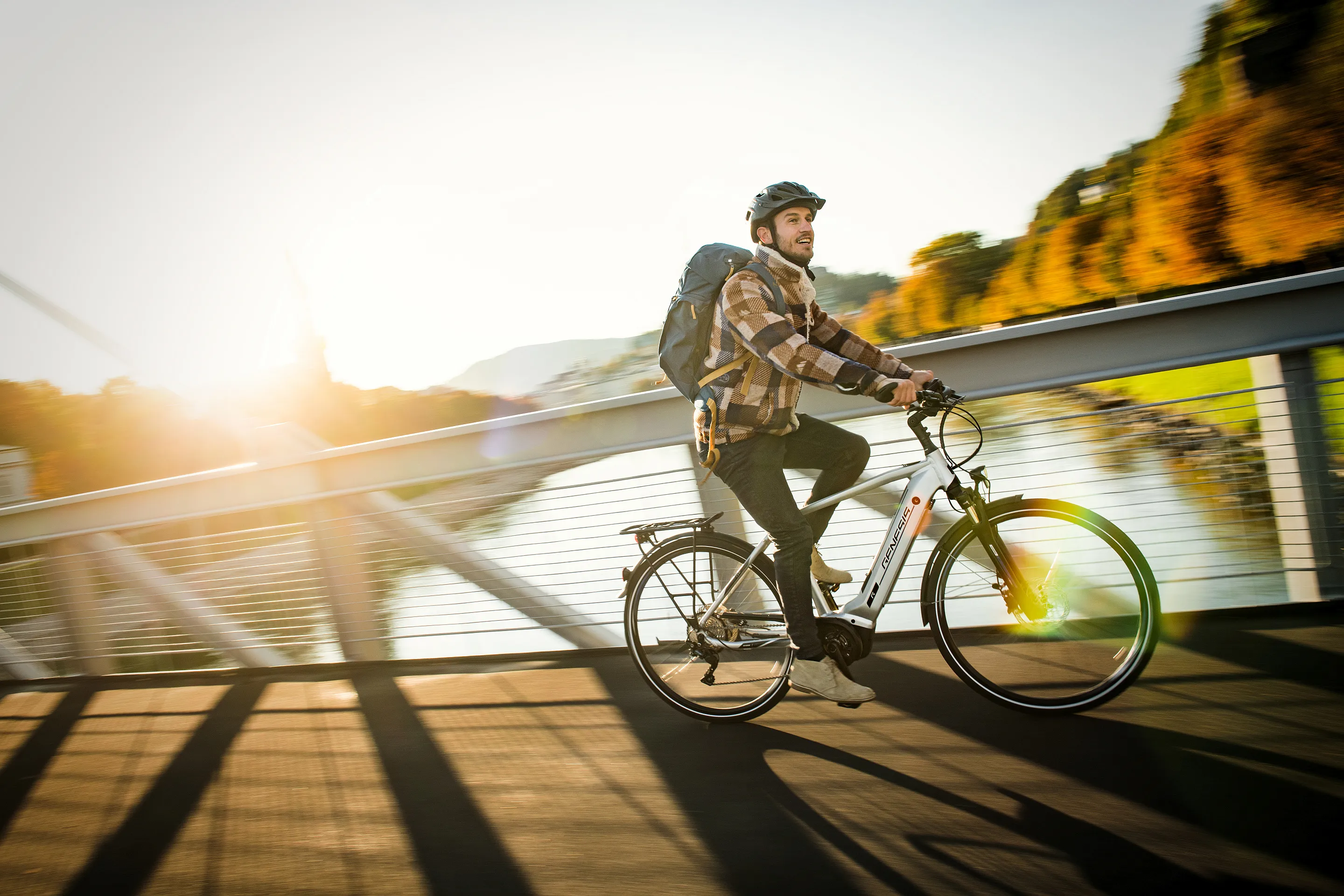 Mann mit Helm und Rucksack fährt bei Sonnenuntergang auf einem weißen Fahrrad über eine Brücke mit Fluss dahinter.