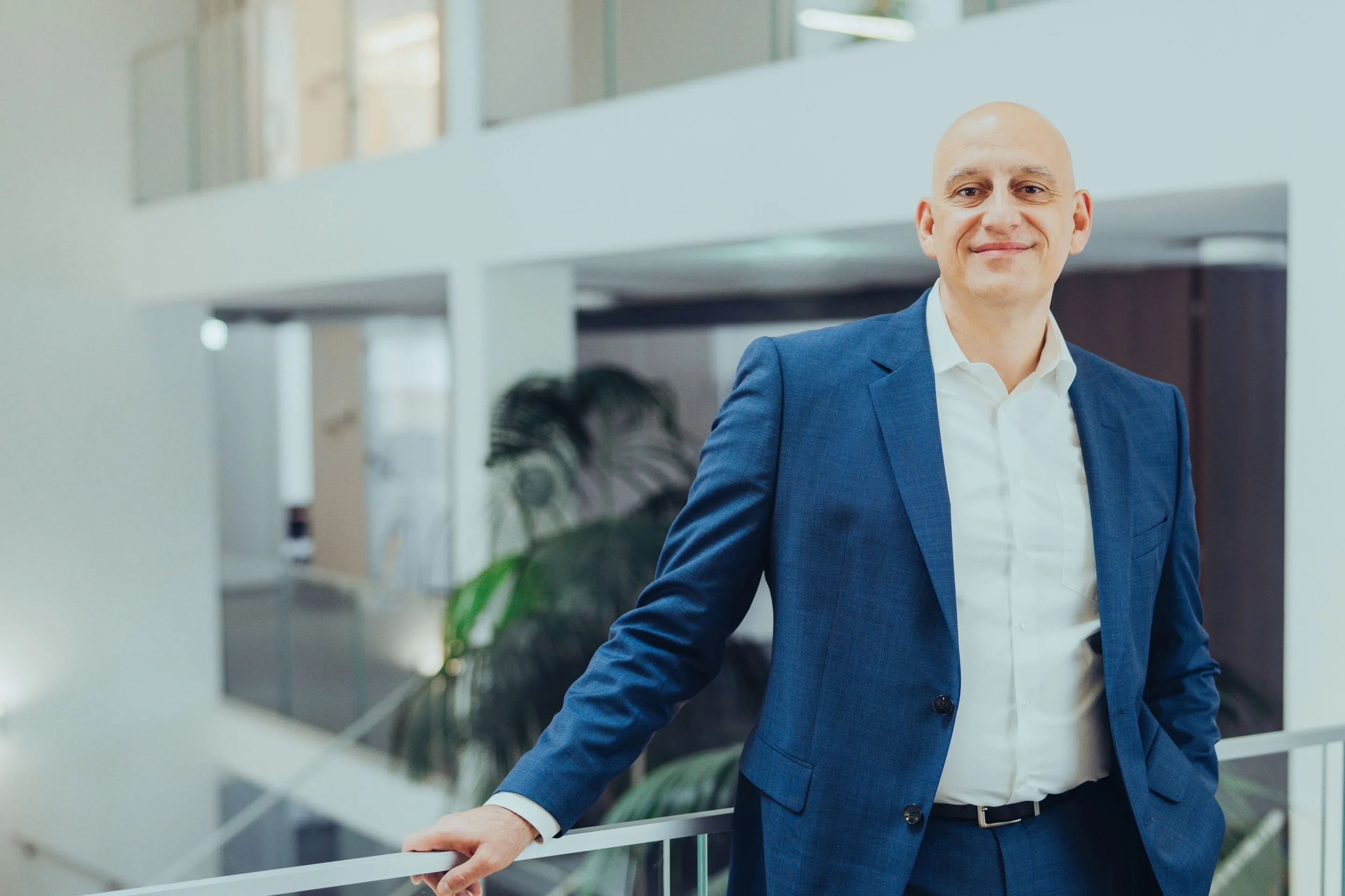 Porträt eines lächelnden, glatzköpfigen Mannes in blauem Anzug und weißem Hemd im modernen Bürogebäude.