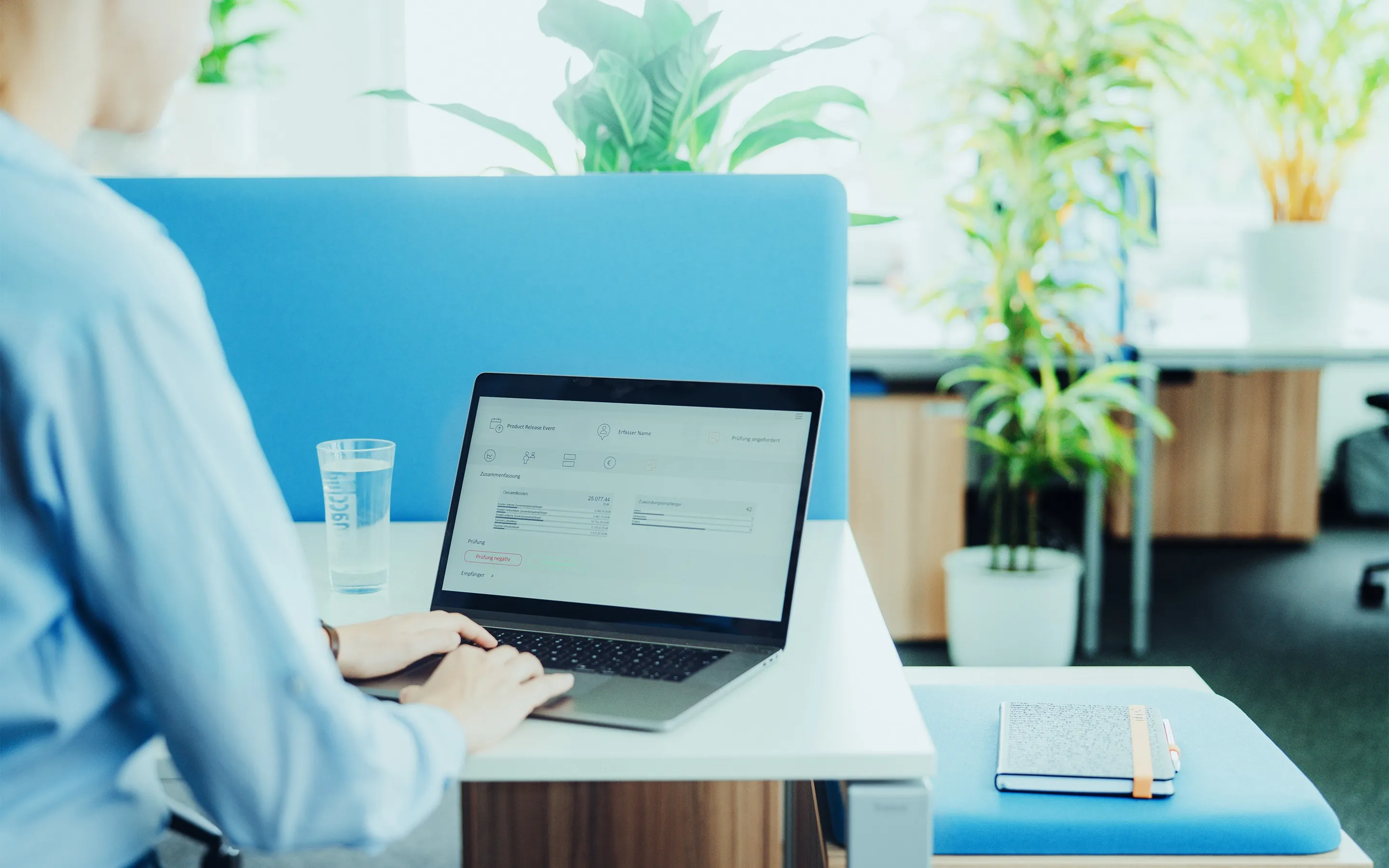 Person arbeitet an einem Laptop an einem hellen Schreibtisch mit Glas Wasser und Notizbuch in einem modernen Büro mit Pflanzen.
