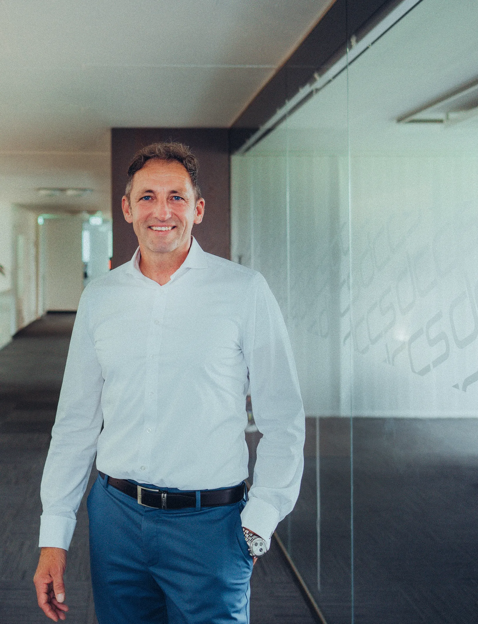 Geschäftsmann in weißem Hemd und blauen Hosen steht lächelnd in einem modernen Büroflur mit Glaswand.