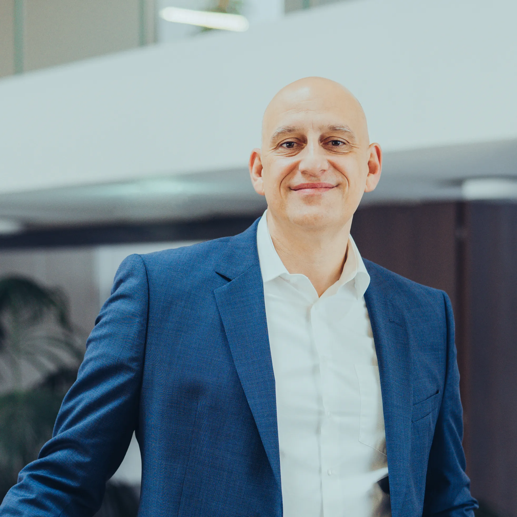 Lächelnder, glatzköpfiger Mann in blauem Anzug und weißem Hemd vor modernem Bürohintergrund.