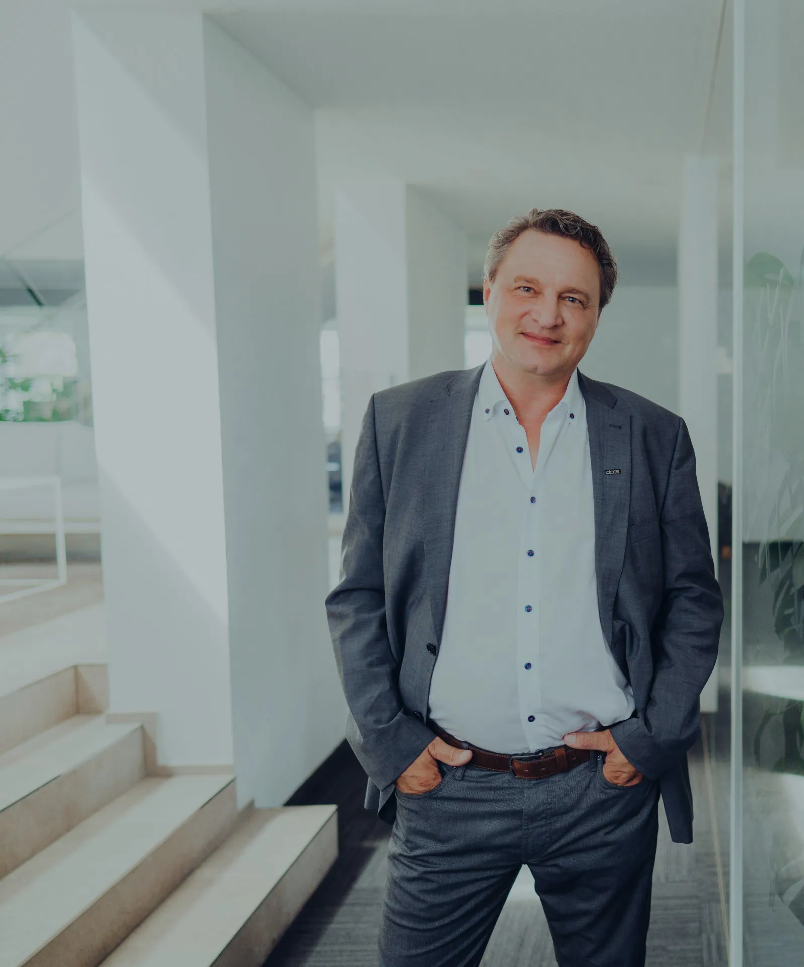 Ein lächelnder Mann in grauem Anzug und weißem Hemd lehnt in einem modernen Büroflur an einer Glaswand.