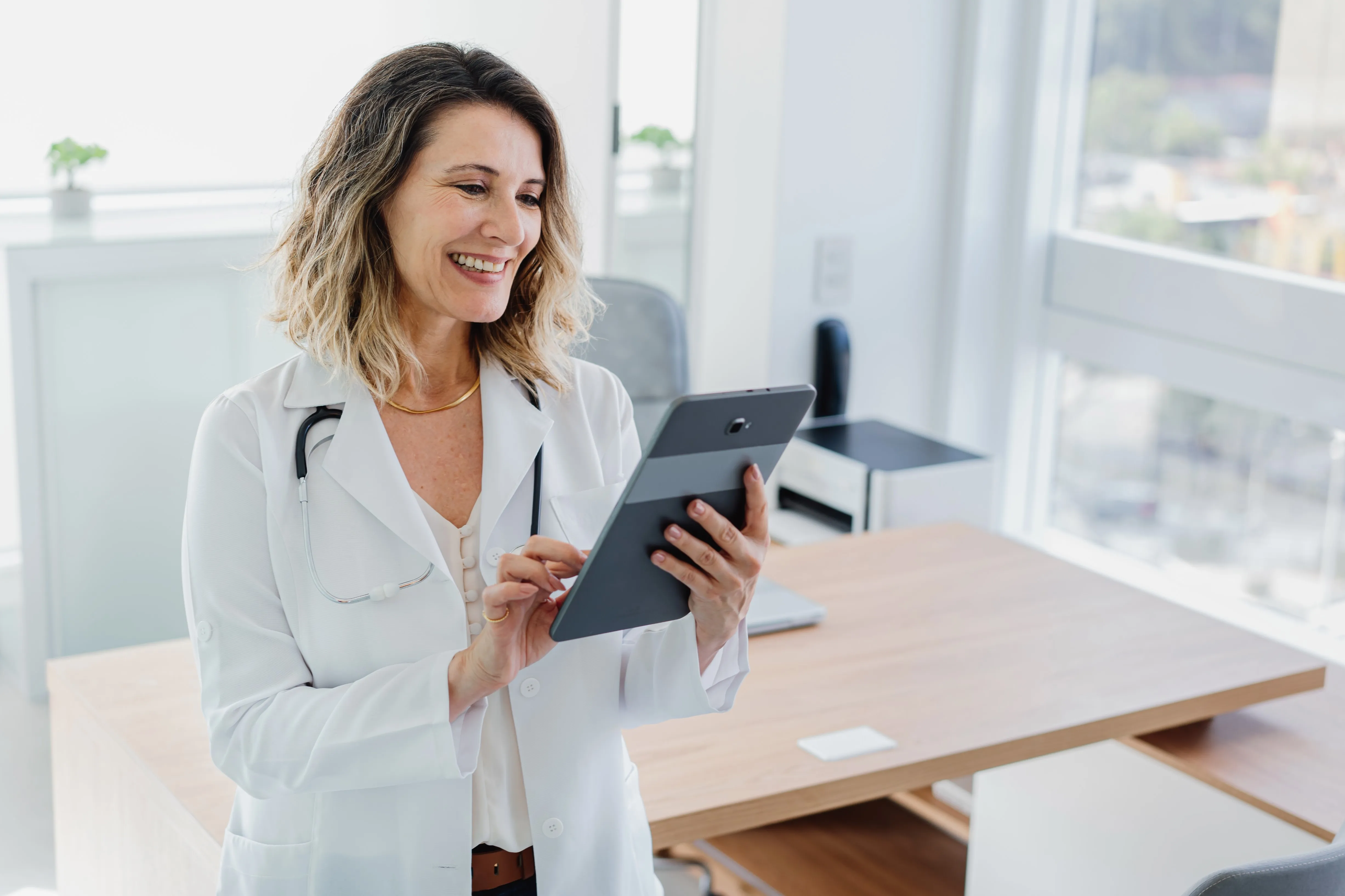 Lächelnde Ärztin mit Stethoskop um den Hals, die ein Tablet in einem hellen Büro benutzt.