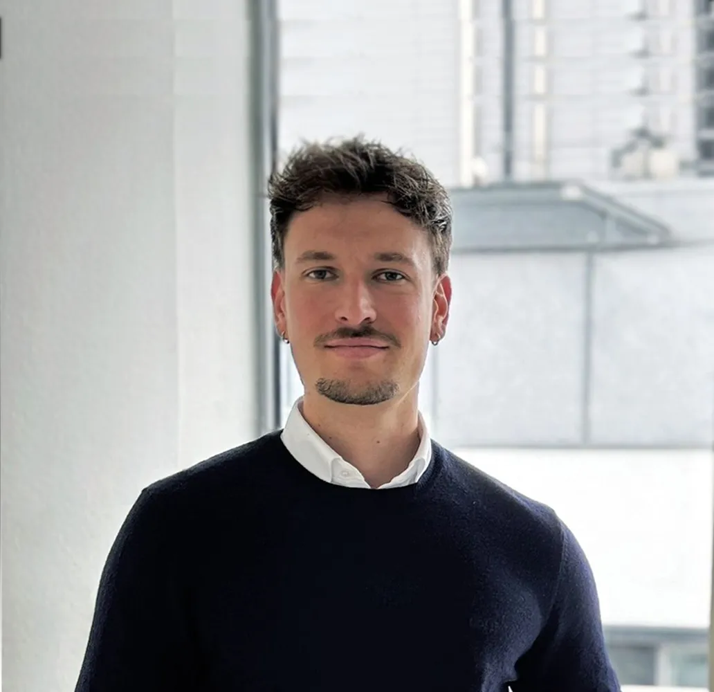 Porträt eines jungen Mannes mit kurzen, gewellten Haaren, leichtem Bart und Ohrsteckern, der einen weißen Hemdkragen unter einem dunklen Pullover trägt, vor einem hellen Fensterhintergrund.