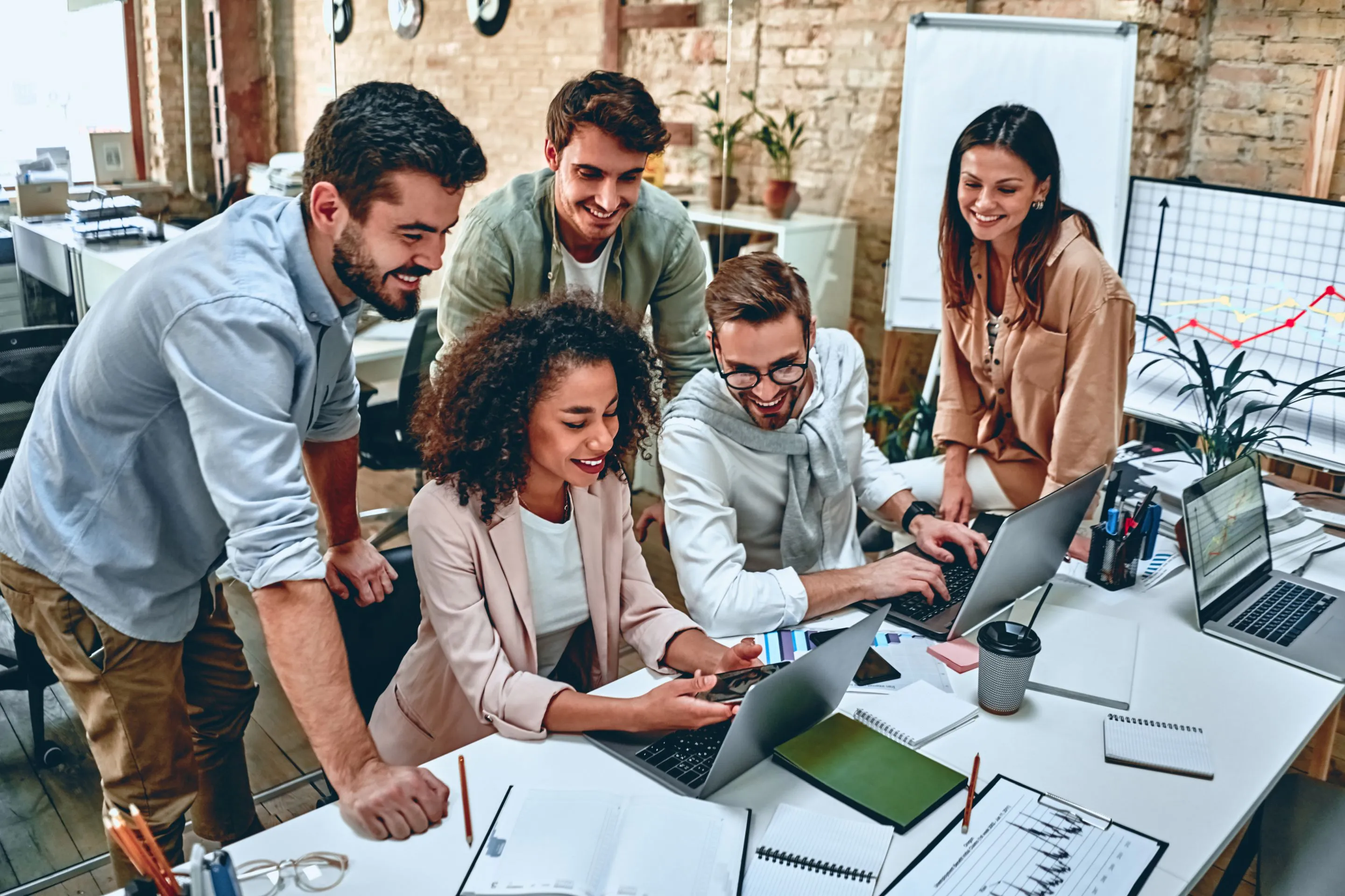 Fünf junge Erwachsene, die in einem modernen Büro an Laptops arbeiten und sich freundlich austauschen.