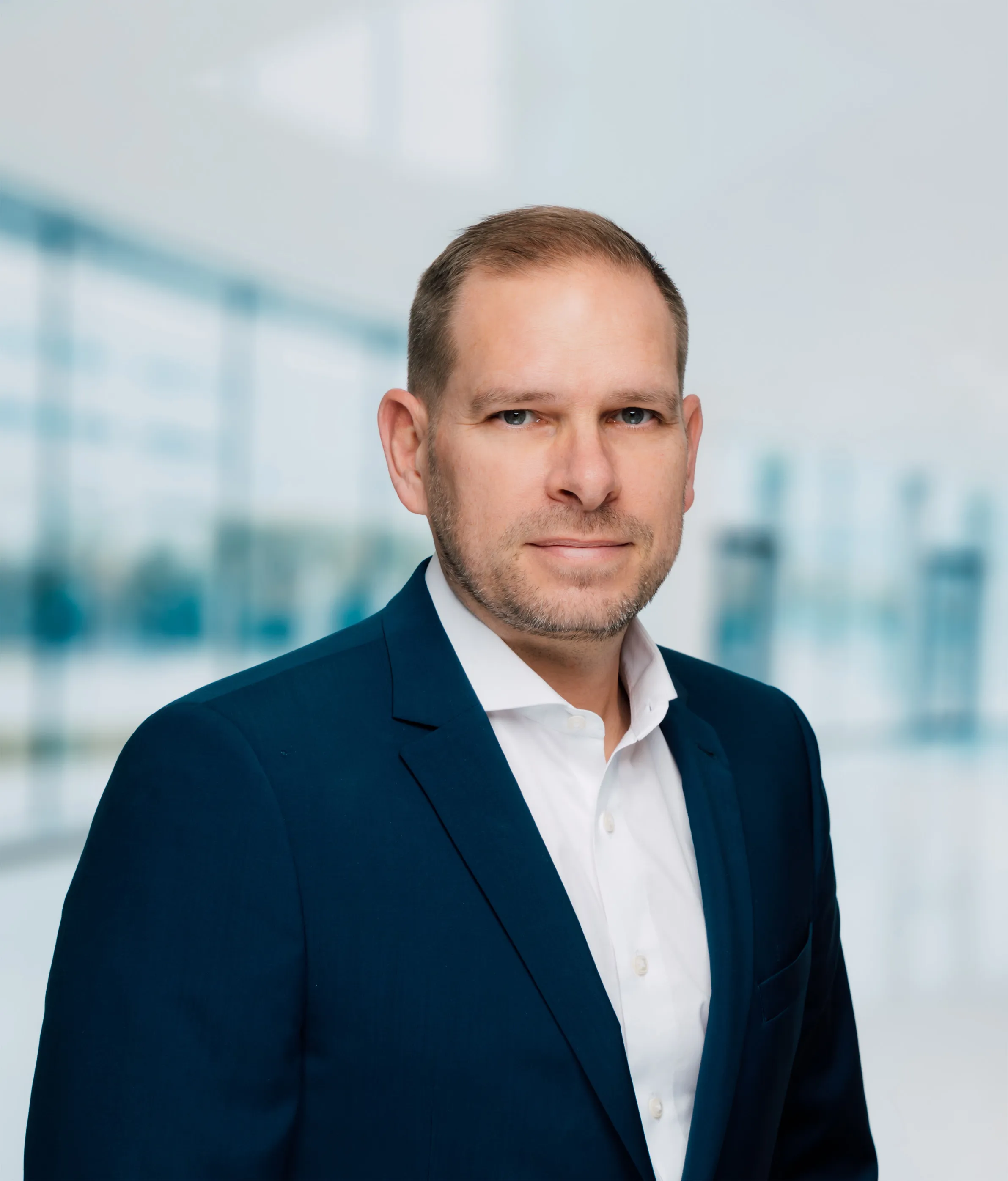 Porträt eines Mannes mit kurzem braunem Haar und Bart, der einen dunkelblauen Anzug und ein weißes Hemd trägt vor einem modernen, hellen Bürohintergrund.
