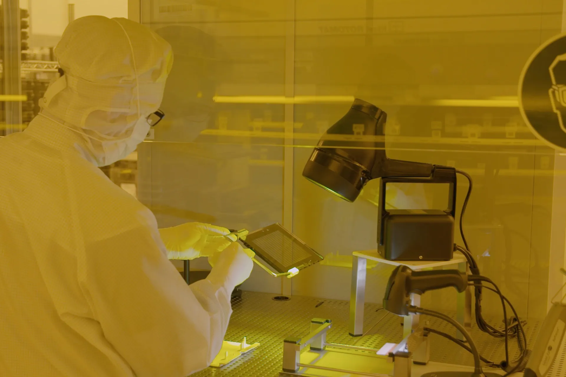 Techniker in Reinraumkleidung untersucht eine elektronische Platte unter gelblichem Licht im Labor.