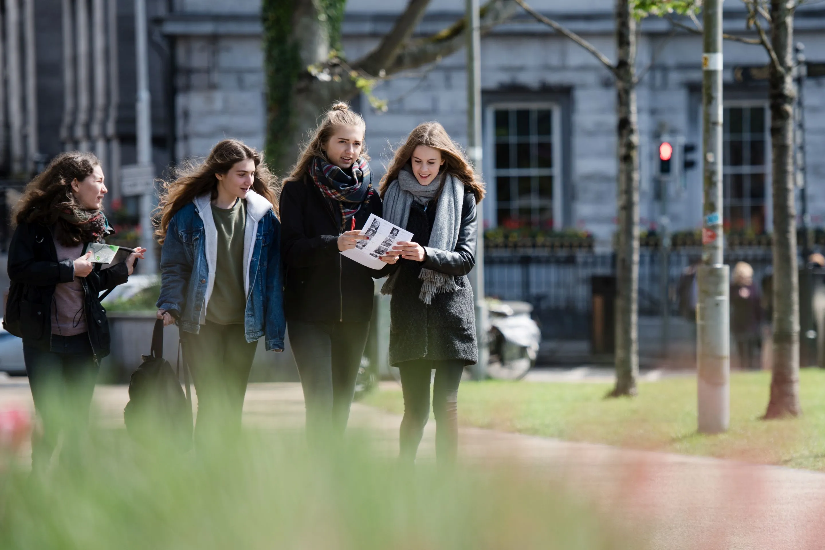 Vier junge Frauen gehen an einem Stadtparkweg, zwei schauen gemeinsam auf ein Blatt Papier.