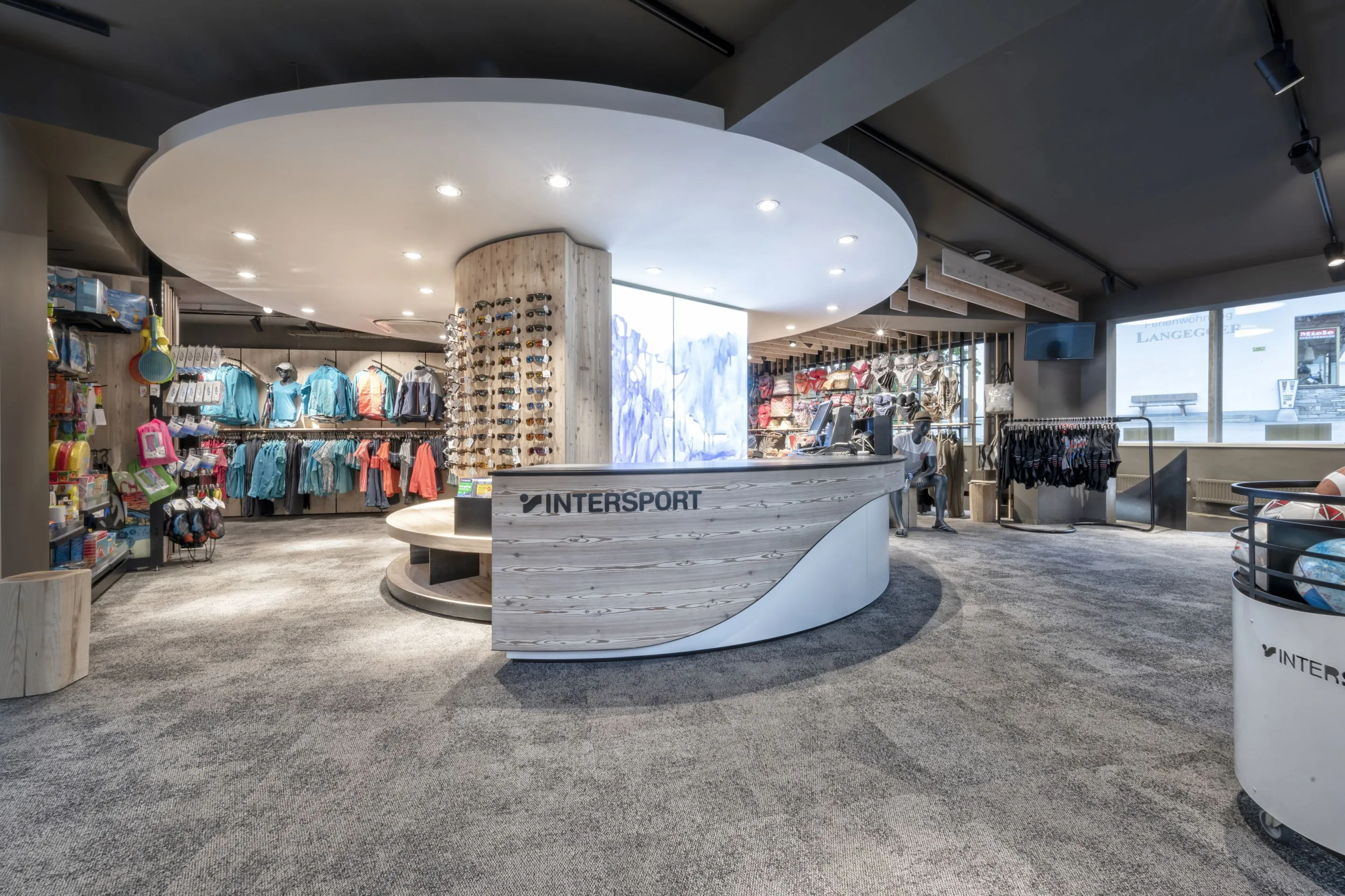 Moderner INTERSPORT-Laden mit eleganter Holztheke, Sonnenbrillenwand, Sportbekleidung und Accessoires auf grauem Teppichboden.