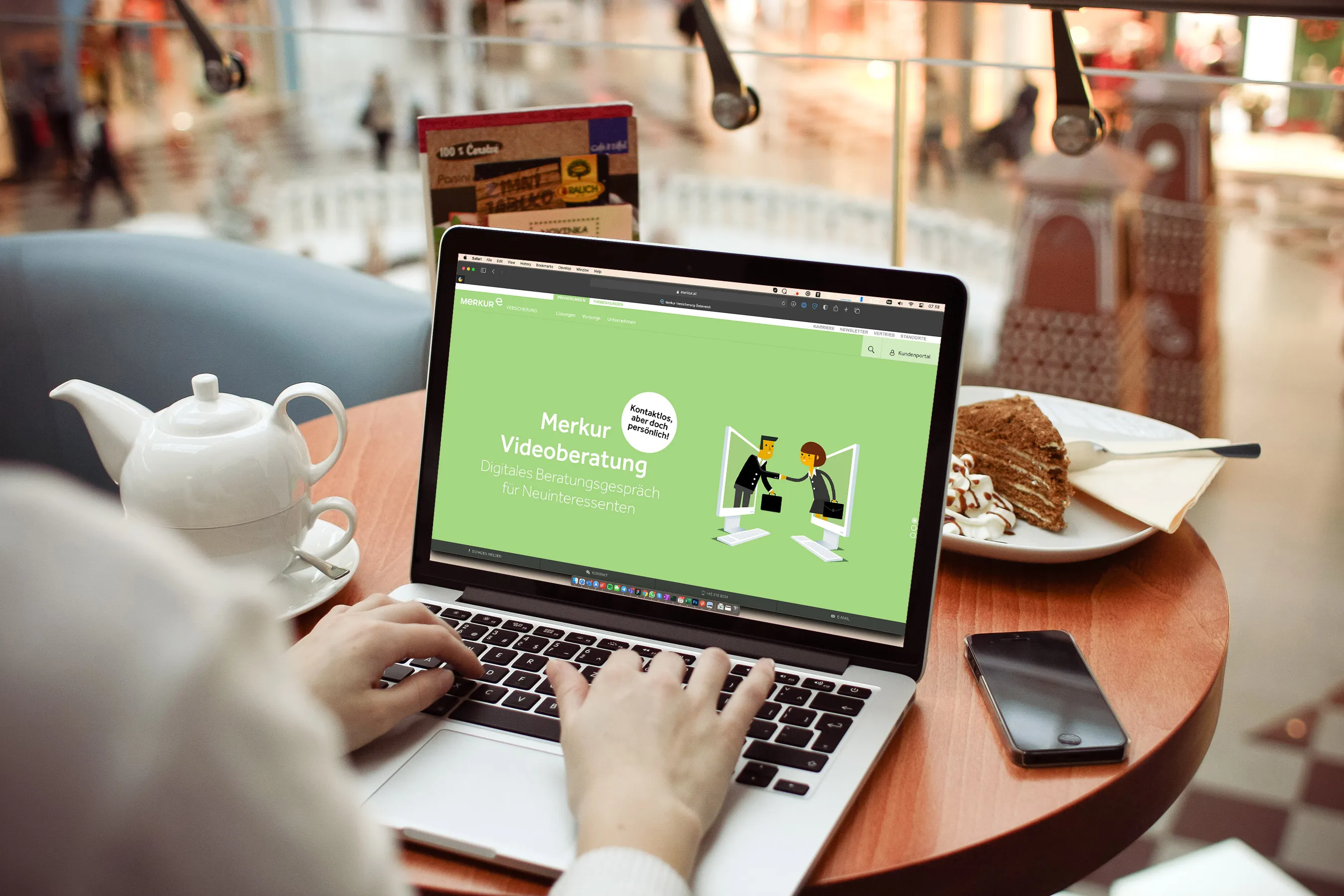 Person tippt auf einem Laptop mit geöffneter Webseite zur Merkur Videoberatung in einem Café, neben einer Tasse Tee, einem Stück Kuchen und einem Smartphone auf dem Tisch.