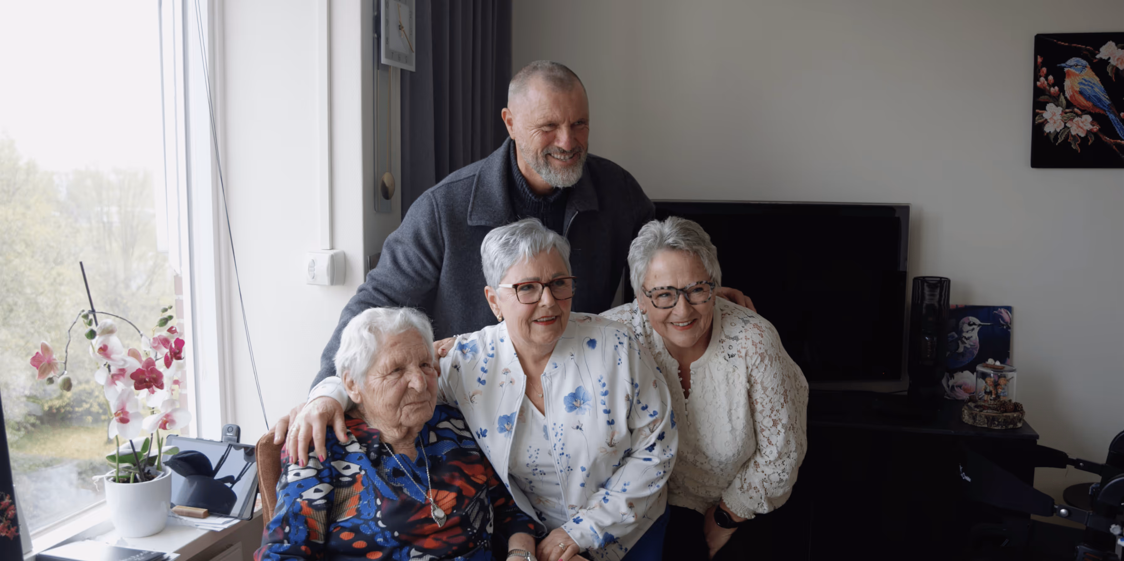 Vier oudere volwassenen, drie vrouwen en een man, lachen samen in een kamer met een raam en planten op de achtergrond.