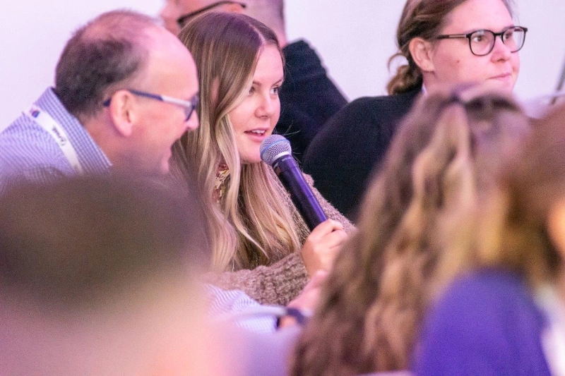 Young woman speaking into a microphone during a group discussion with other attentive people around her.
