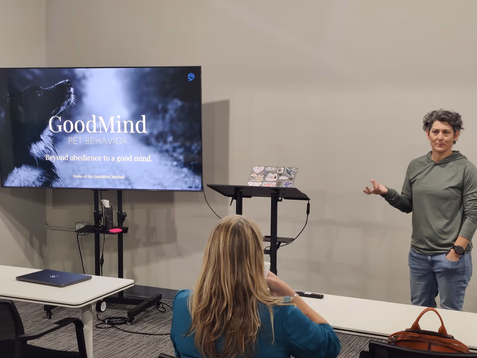 A woman in a green hoodie gives a presentation on GoodMind Pet Behavior to another woman seated at a table, with a laptop and a screen displaying a dog and presentation title.