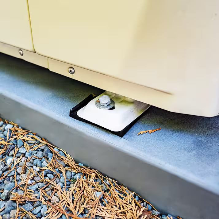 Close-up of a metal mounting bracket with a bolt securing the base of a beige outdoor unit on a gray metal platform surrounded by gravel and dried pine needles.