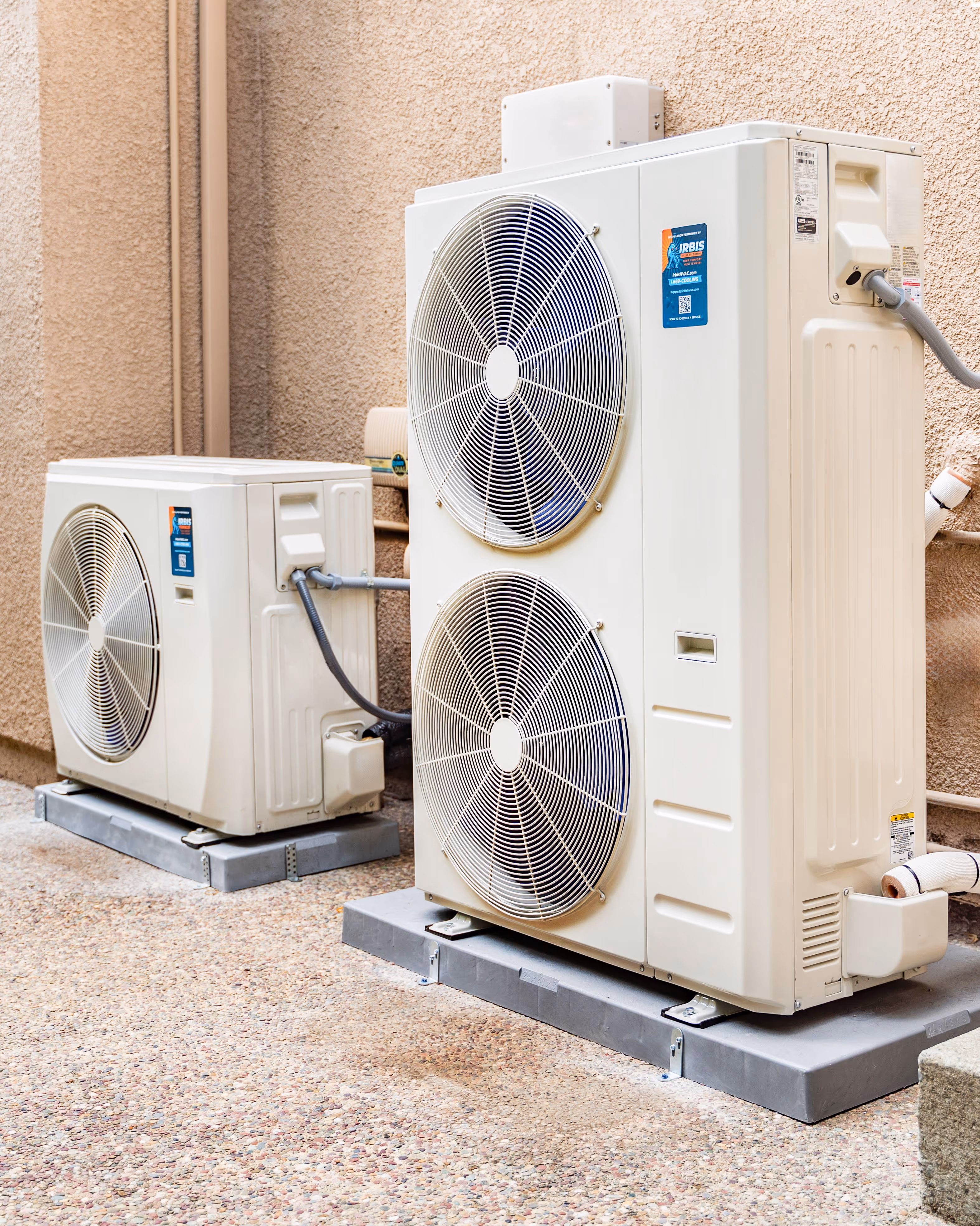 Two white outdoor air conditioning units with large circular fans mounted on concrete pads beside a beige stucco wall.