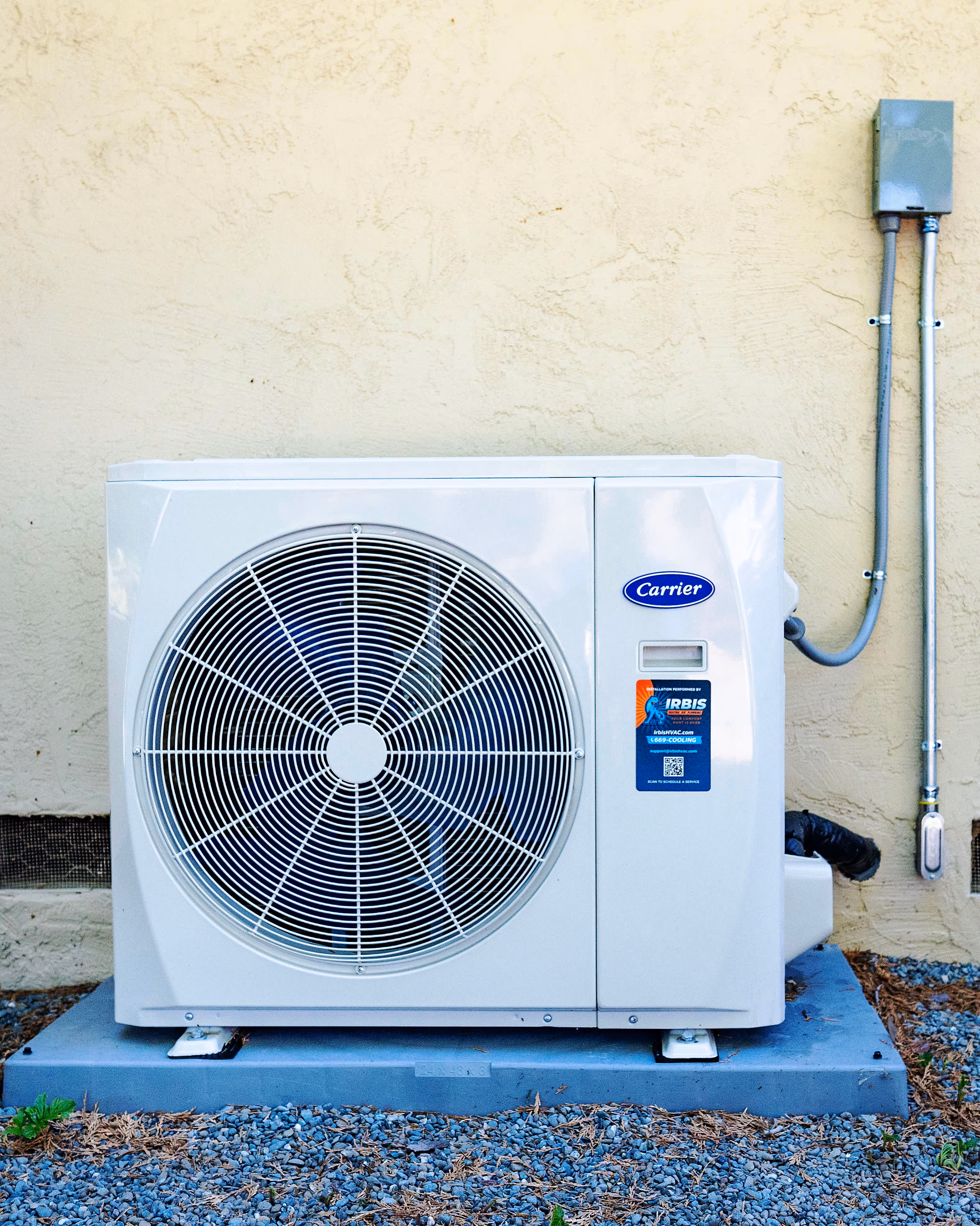 Outdoor air conditioning unit mounted on a concrete slab against a beige stucco wall with an electrical junction box and conduit.