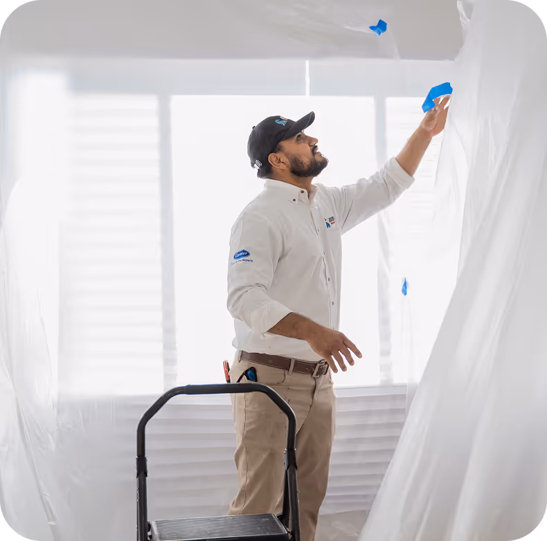 Technician in uniform taping plastic sheeting to cover a window for protection during work.