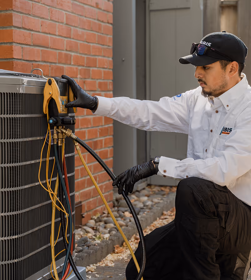 Technician wearing black gloves and uniform testing an outdoor air conditioning unit with gauges and hoses.