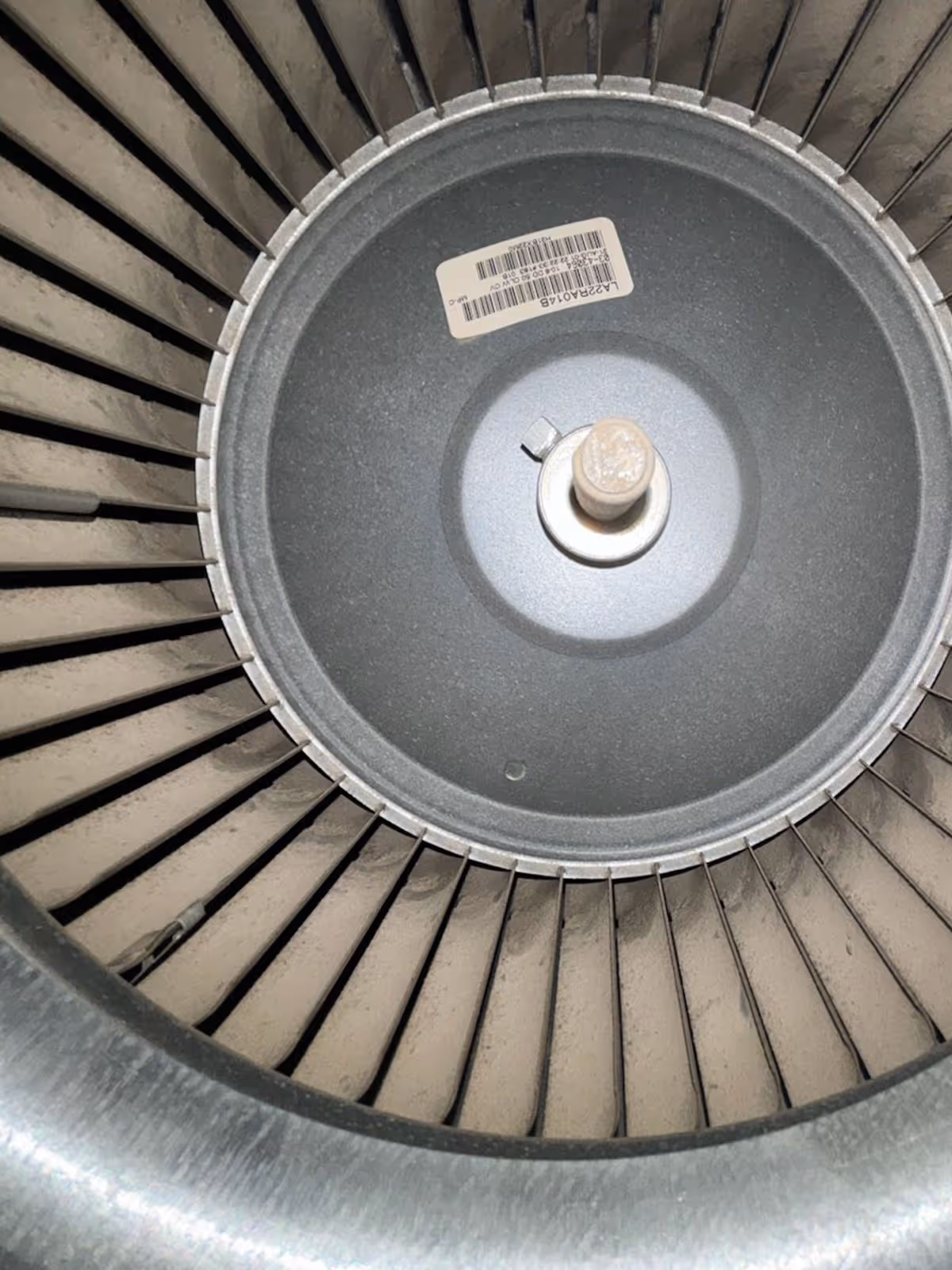 Close-up of a dusty circular fan or blower wheel with metal blades and a central mounting hub with a barcode sticker.