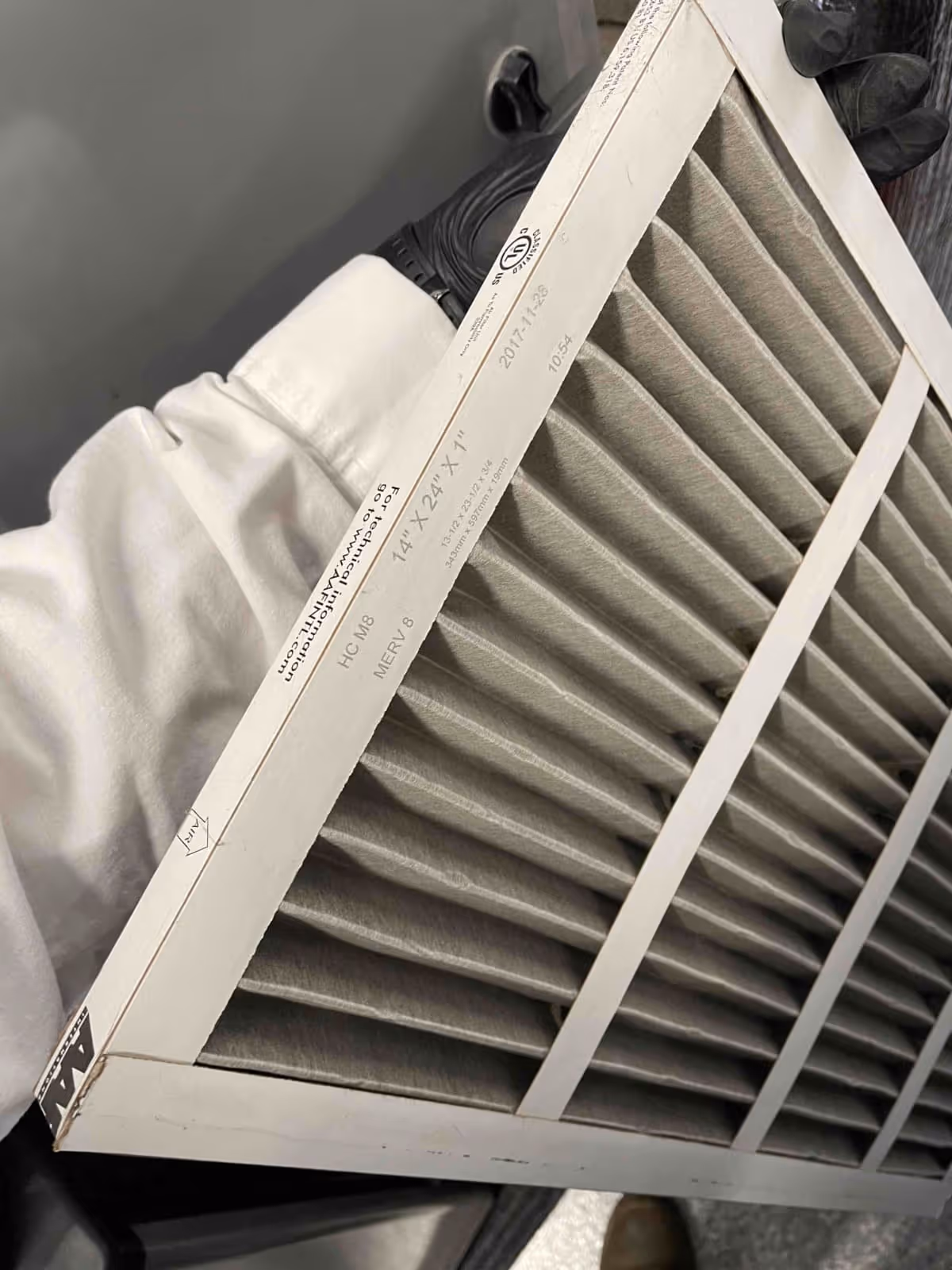 Person wearing black gloves holding a rectangular 14x24x1 inch HVAC air filter with white frame and pleated fiberglass material.
