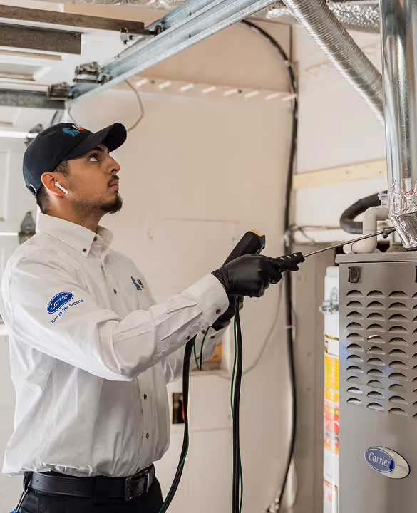 Technician in white uniform and black cap inspecting a furnace with a diagnostic tool.