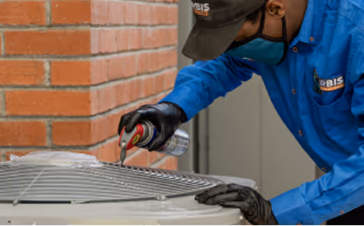 Technician in blue uniform cleaning an outdoor air conditioning unit with a spray can.