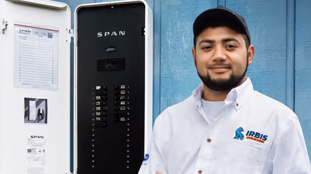 Technician in white shirt and cap standing beside an open SPAN electrical panel on blue wall.