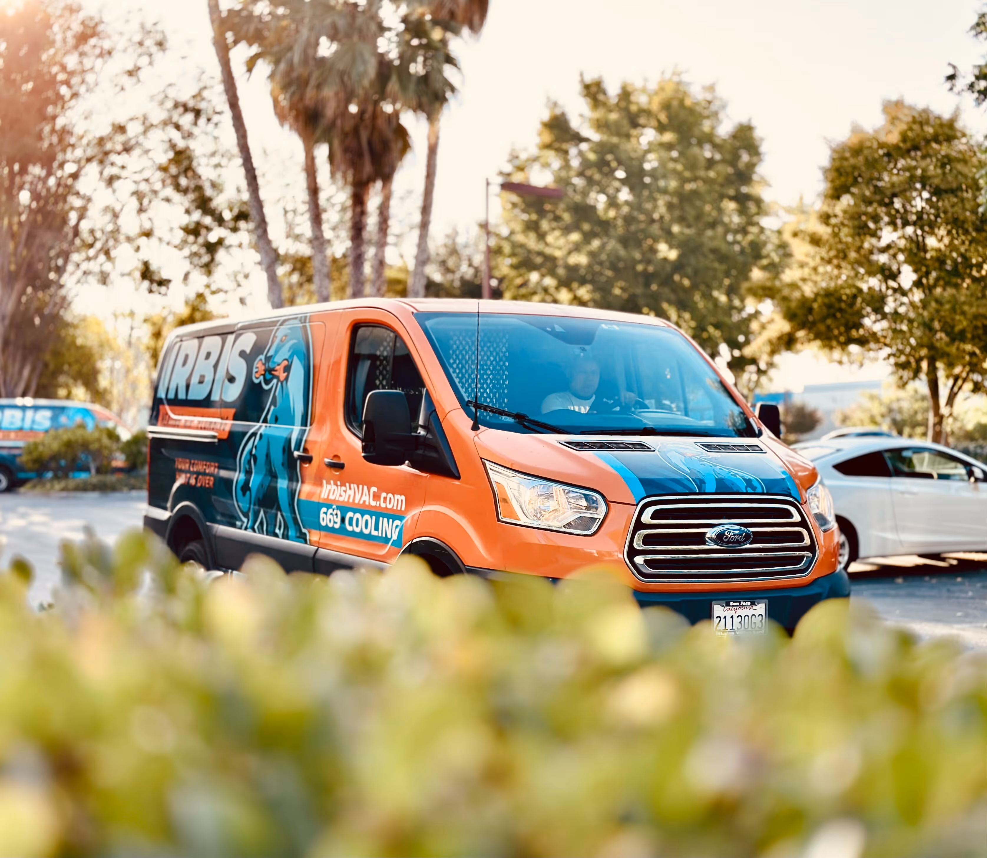 Orange and blue Irbis HVAC service van parked outdoors with palm trees and greenery in background.