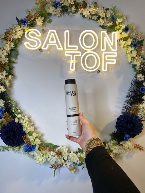 Een hand houdt een fles conditioner voor een verlicht neonbord met de tekst 'SALON TOF' omringd door een bloemenkrans aan de muur van een kapperszaak in 's-Hertogenbosch.