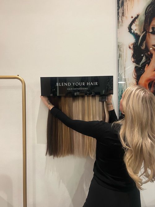 Een vrouwelijke kapper in 's-Hertogenbosch plaatst een paneel met voorbeelden van blonde en bruine hairextensions aan de muur in een moderne kapsalon.