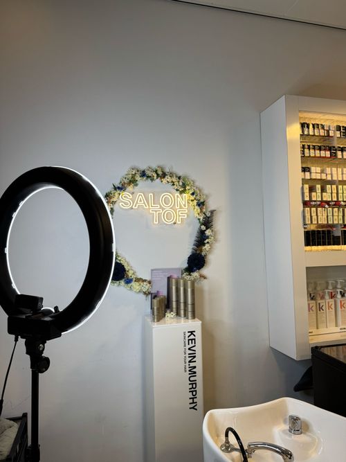 Interieur van een kapperszaak in 's-Hertogenbosch met een verlichte ringlamp, een wasbak, en een muurdecoratie dat "SALON TOF" omringd door bloemen leest.