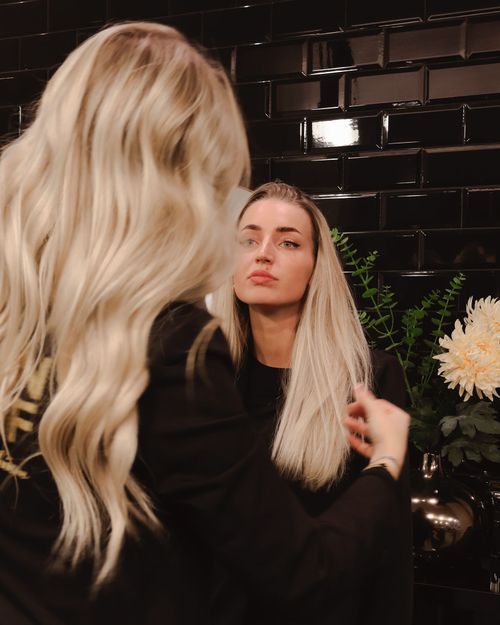 Een vrouw met lang blond haar bekijkt haar reflectie in een spiegel in een kapperszaak in 's-Hertogenbosch, met een achtergrond van zwarte tegels en een vaas met bloemen.