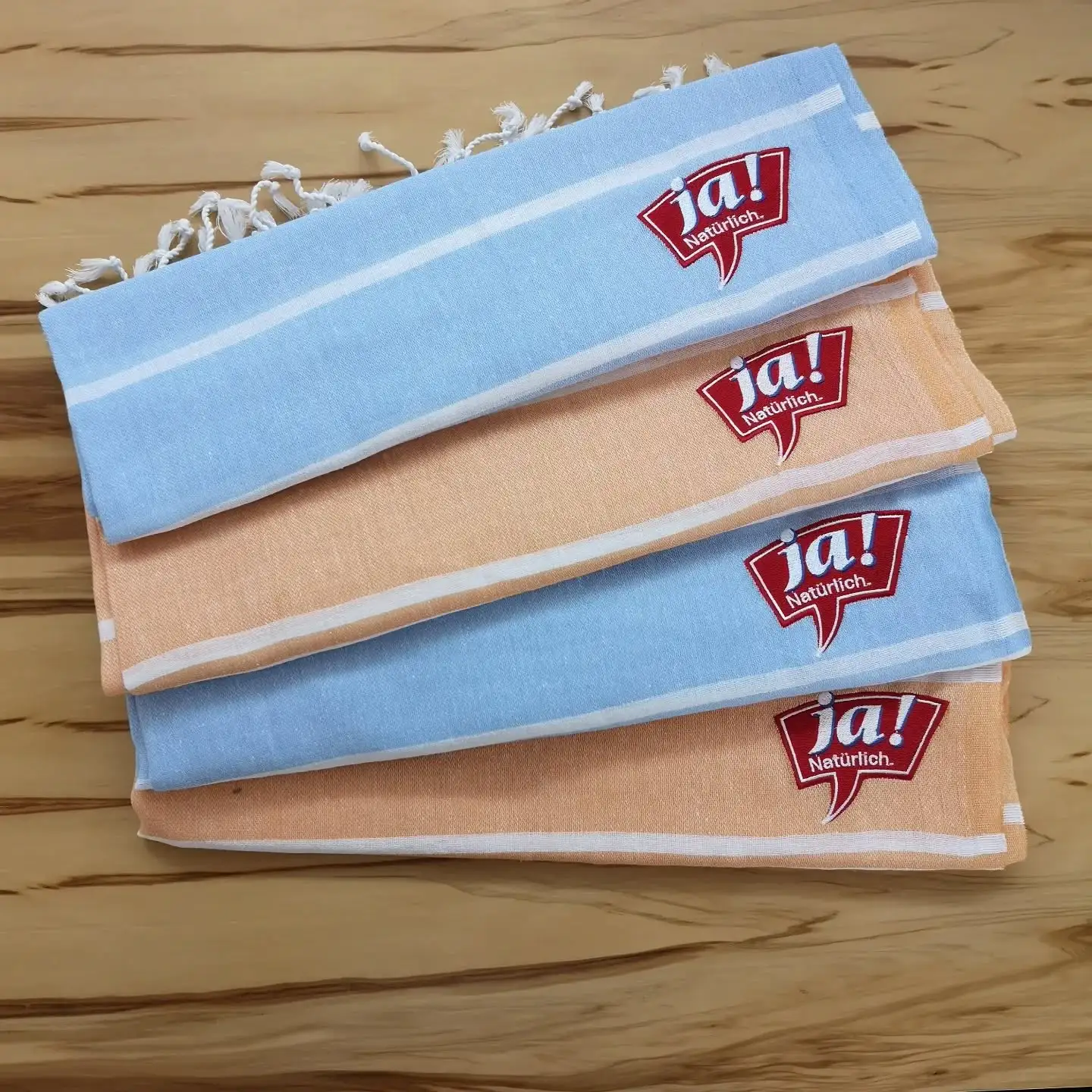 Gestapelte Tücher mit „ja! natürlich“-Stickerei in Blau und Orange.