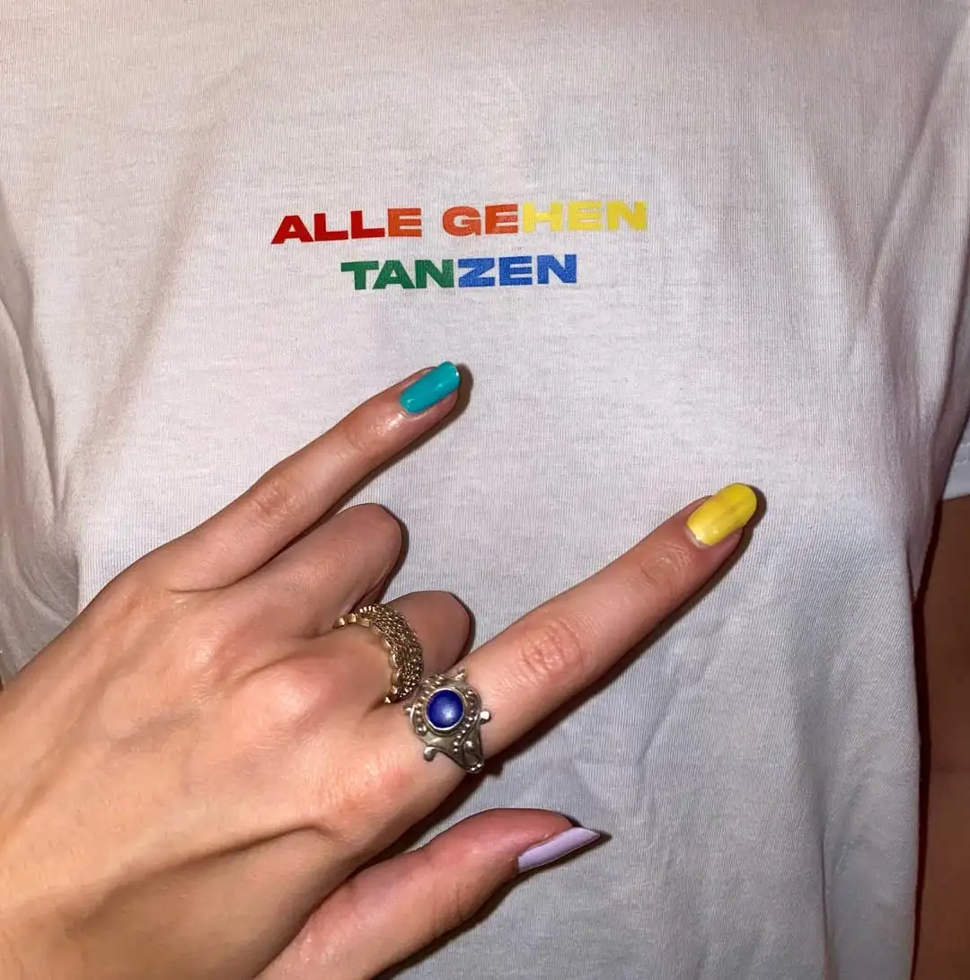 Hand zeigt auf weißes T-Shirt mit bunter Stickerei „ALLE GEHEN TANZEN“.