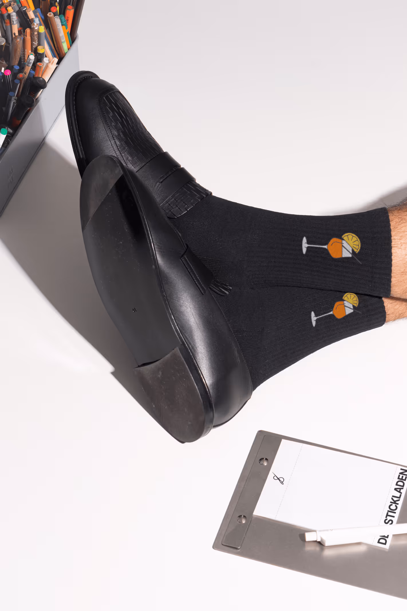 Schwarze Socken mit Cocktail-Stickerei, getragen mit schwarzen Lederschuhen, neben Klemmbrett und Stiften.