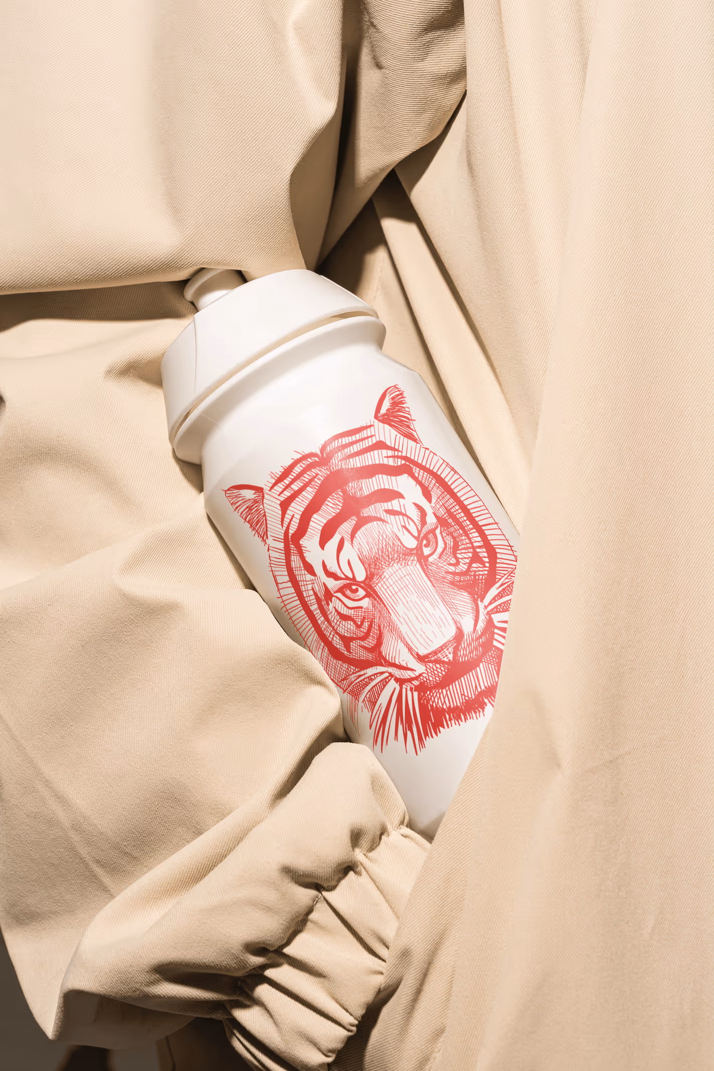 Weisse Trinkflasche mit rotem Tigerkopf-Motiv wird in beigem Mantel gehalten.