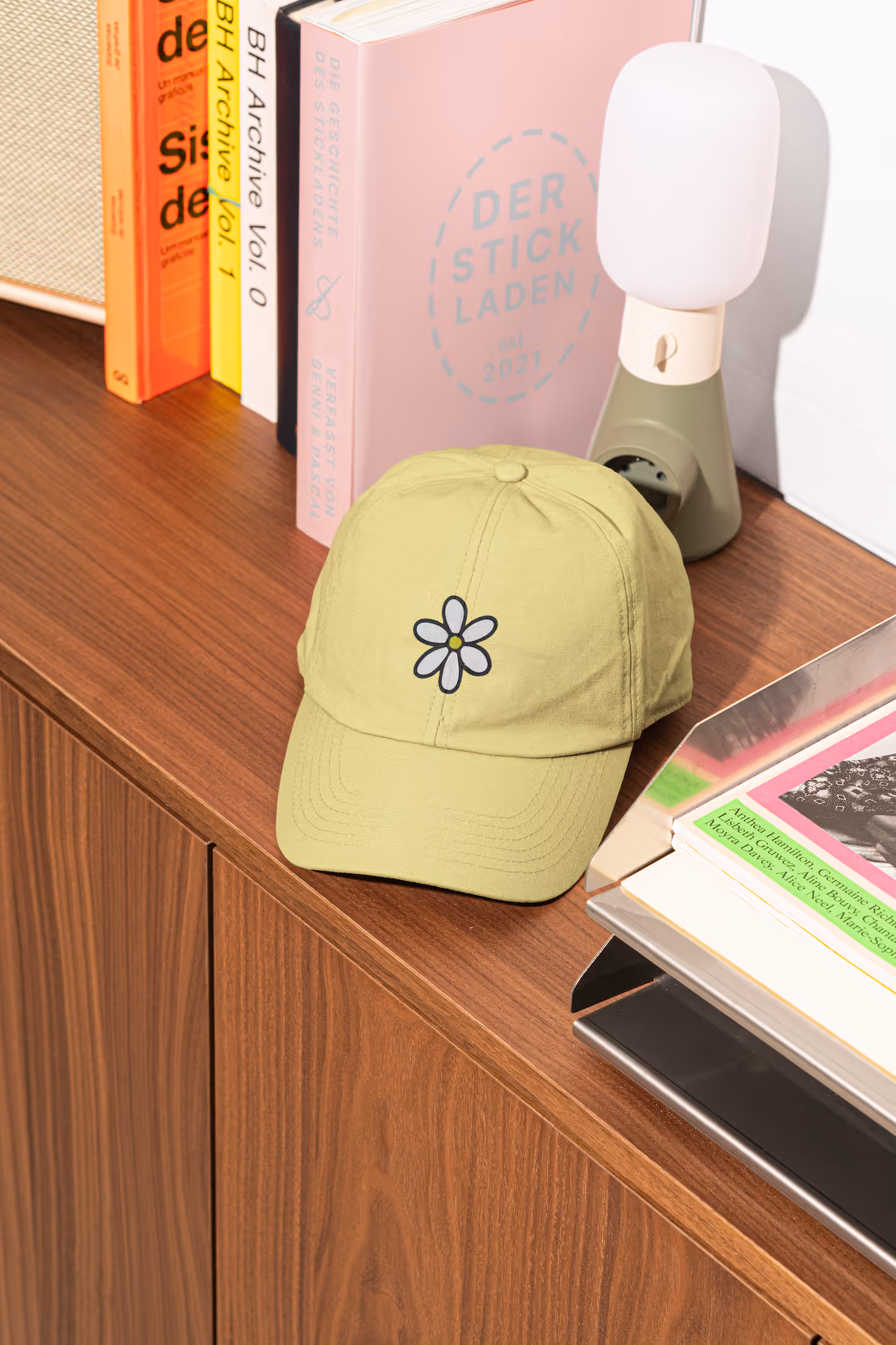 Gelbe Baseballkappe mit weisser Blume liegt auf Holzkommode neben Büchern und Lampe.