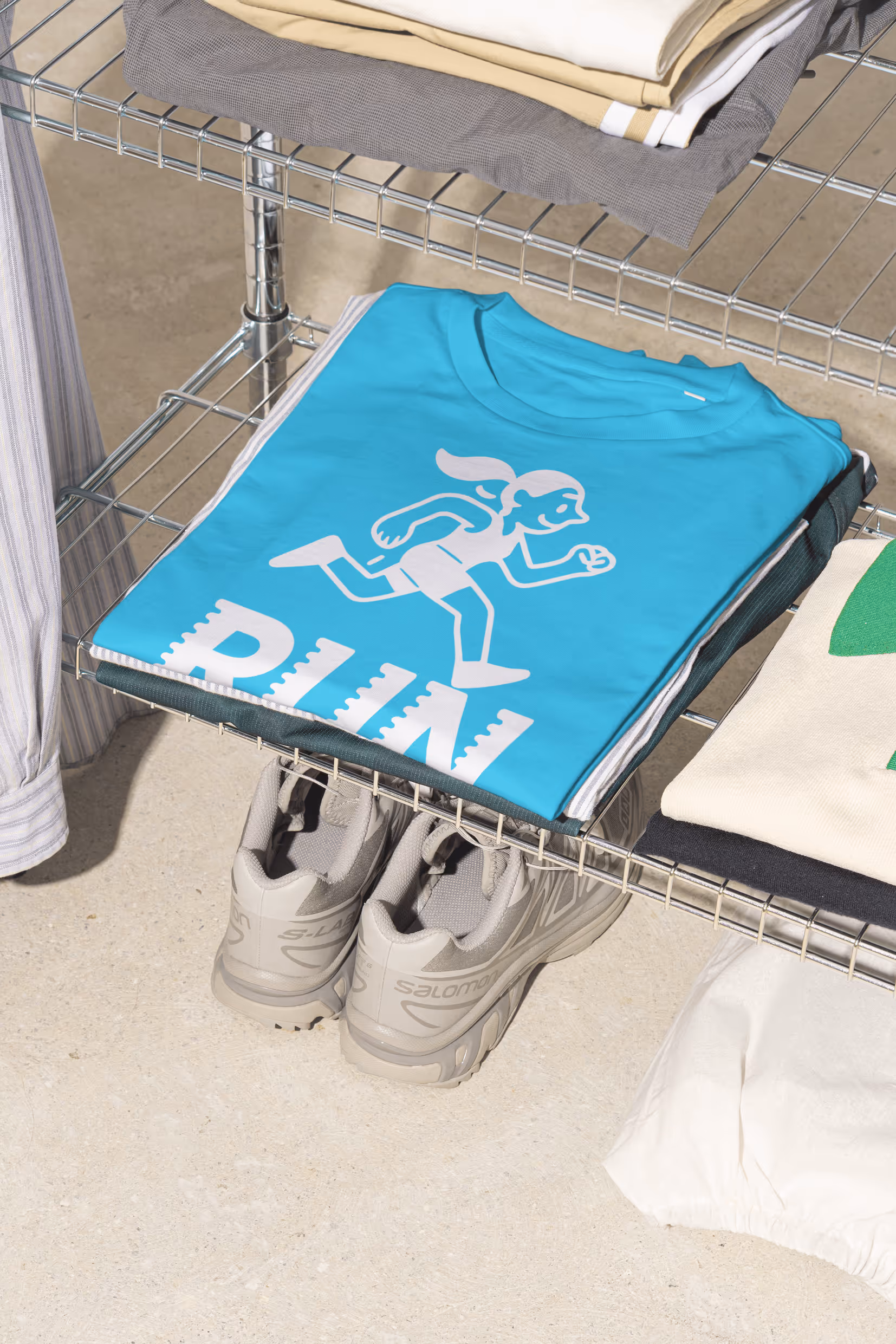 Gefaltetes hellblaues T-Shirt mit laufender Figur und „RUN“-Schriftzug in Metallregal, darunter graue Sneakers.