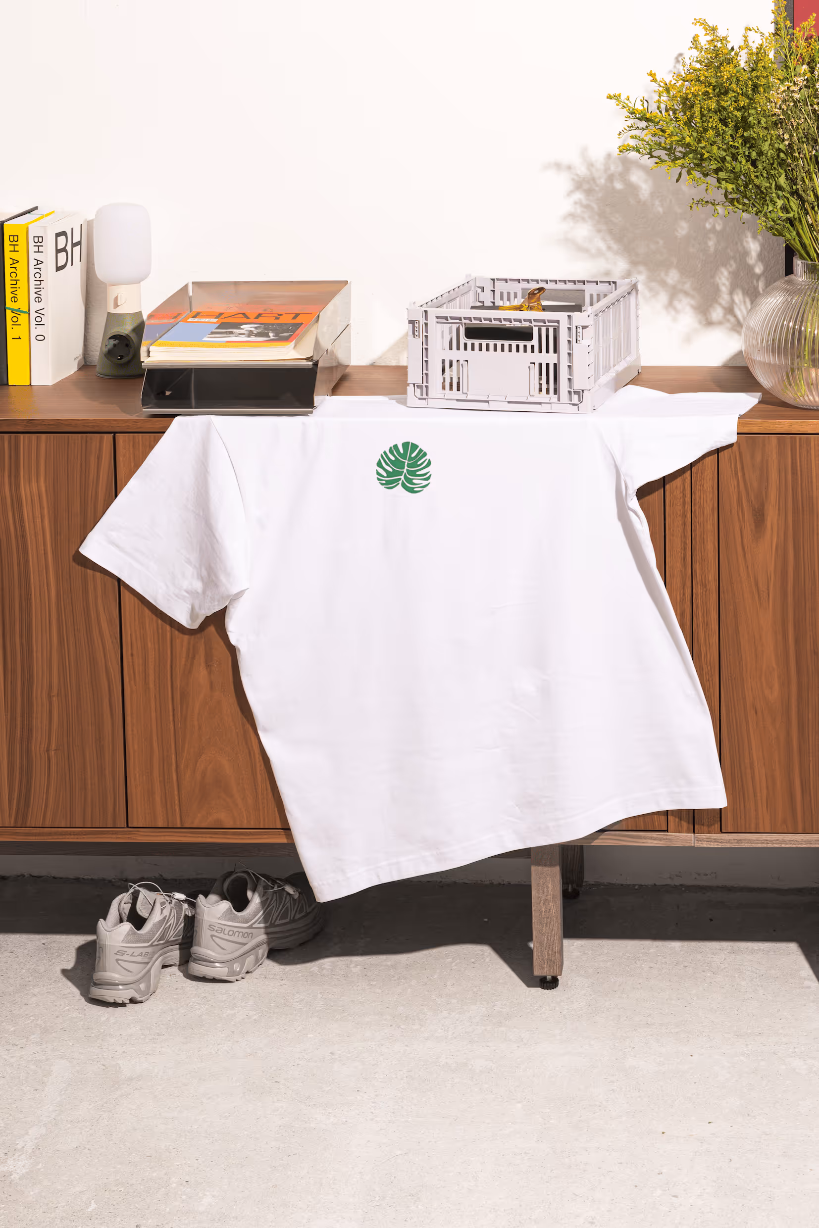 Weisses T-Shirt mit grünem Monstera-Blatt liegt locker auf Holzkommode, darunter ein Paar graue Sneakers.
