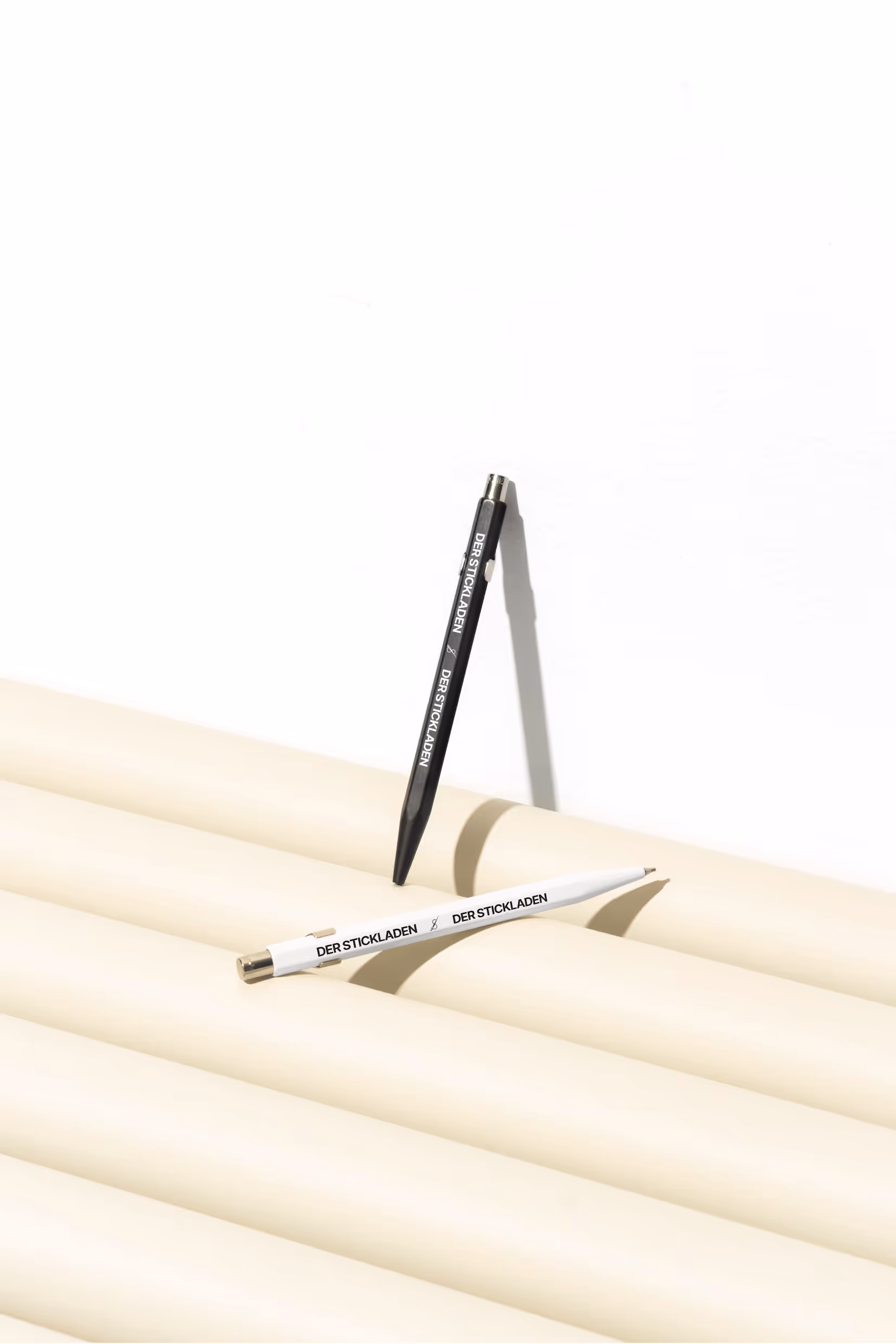 Zwei Kugelschreiber mit „DER STICKLADEN“-Aufdruck auf geriffelter, heller Oberfläche.